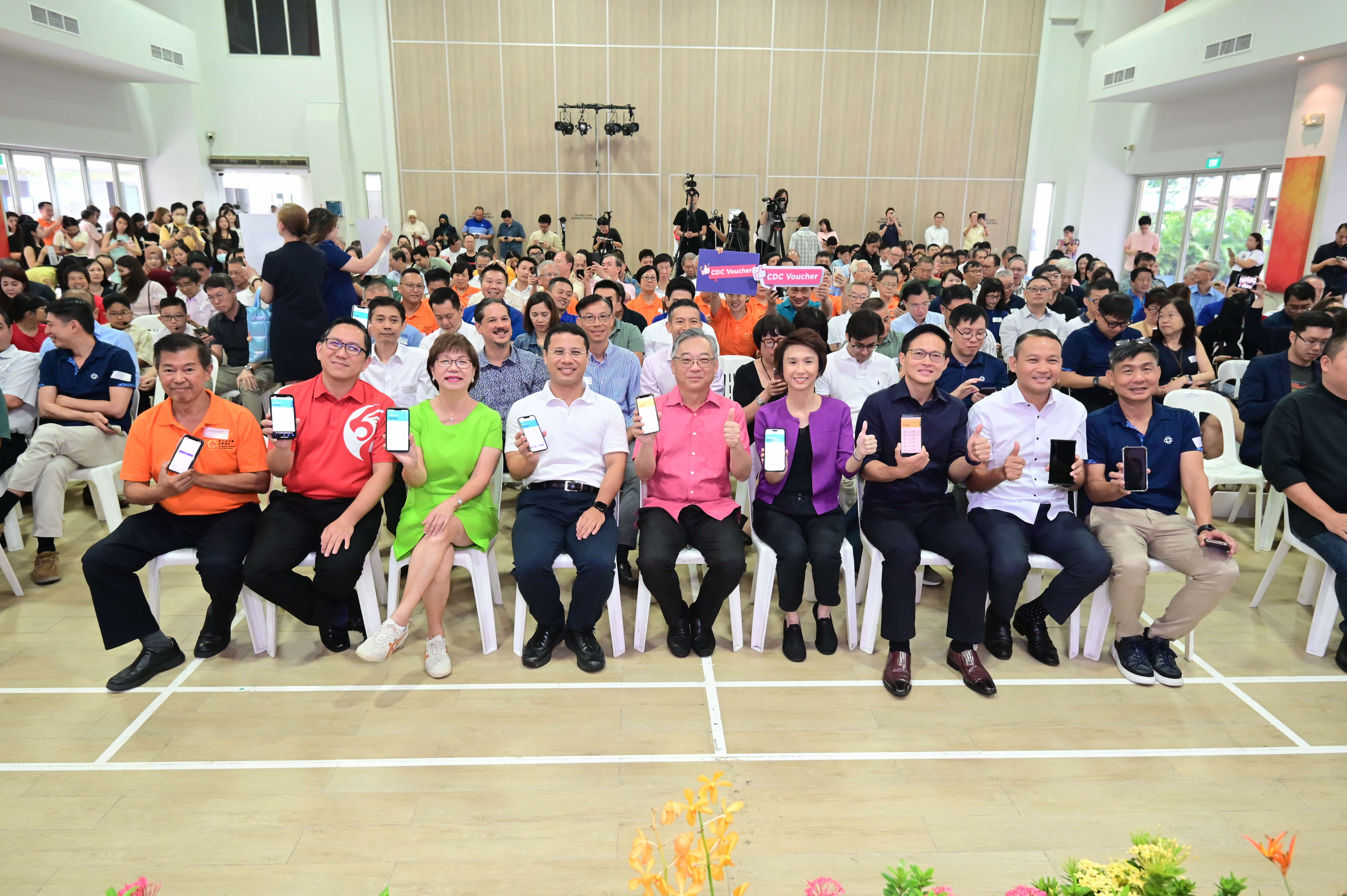 A large group of people seated in rows in a hall, many holding up phones displaying various screens.