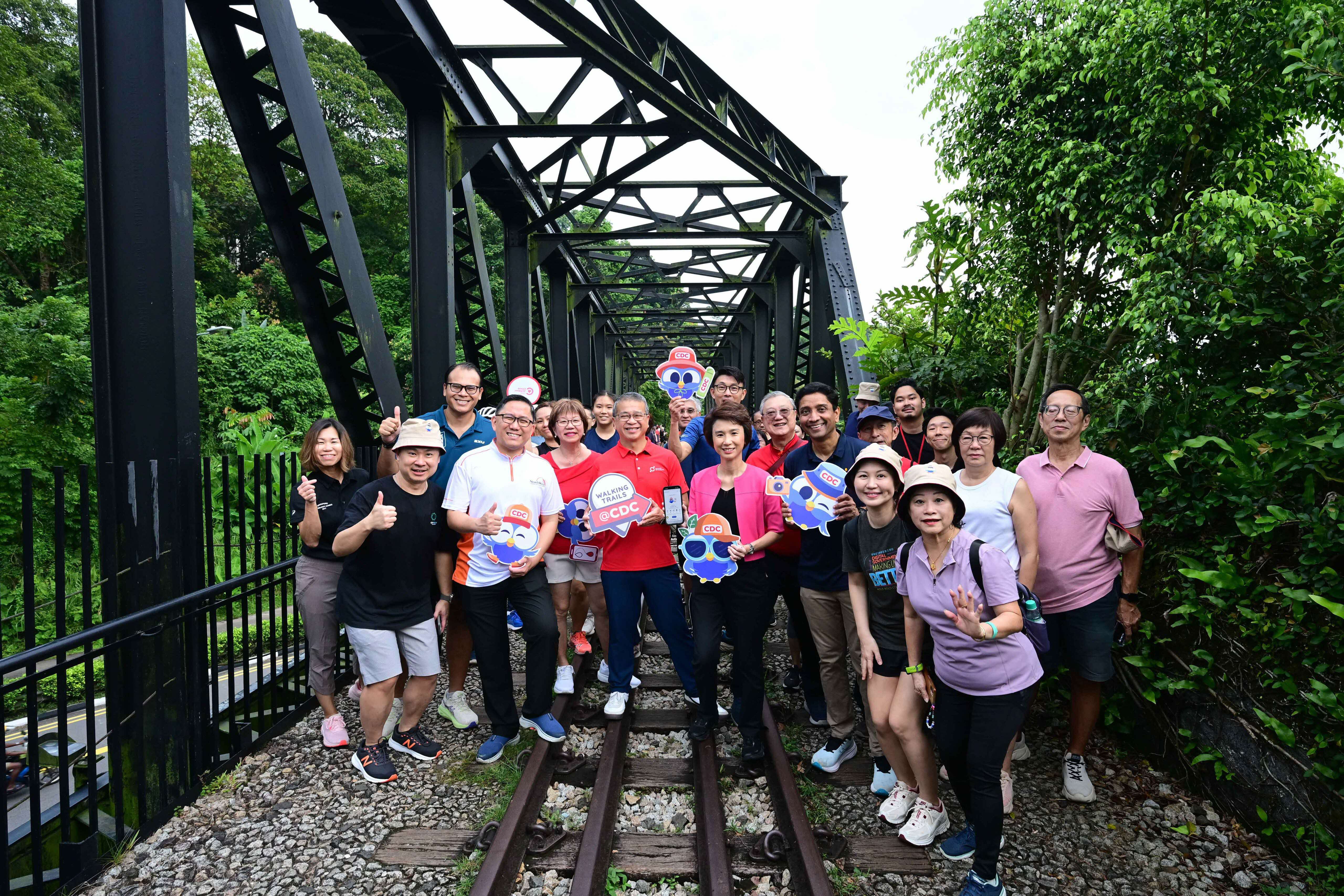 A group of people standing on a railway track under a metal bridge surrounded by lush greenery.