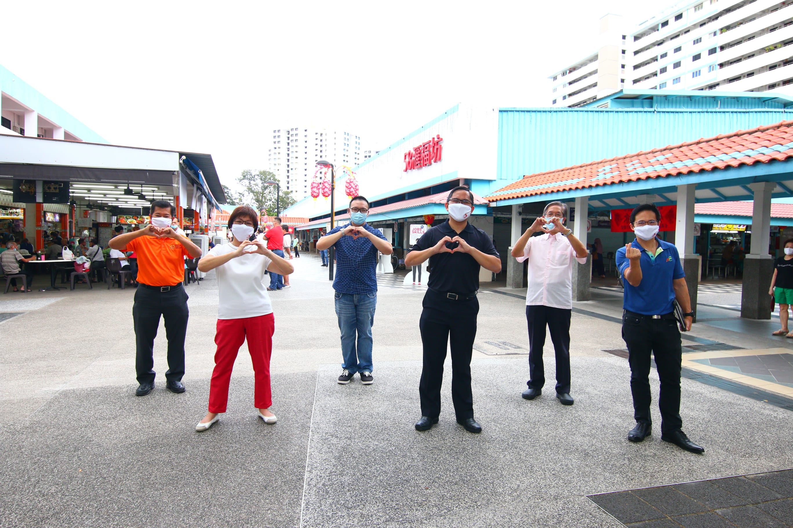 Six people standing in front of a hawker center making heart shapes with their hands.