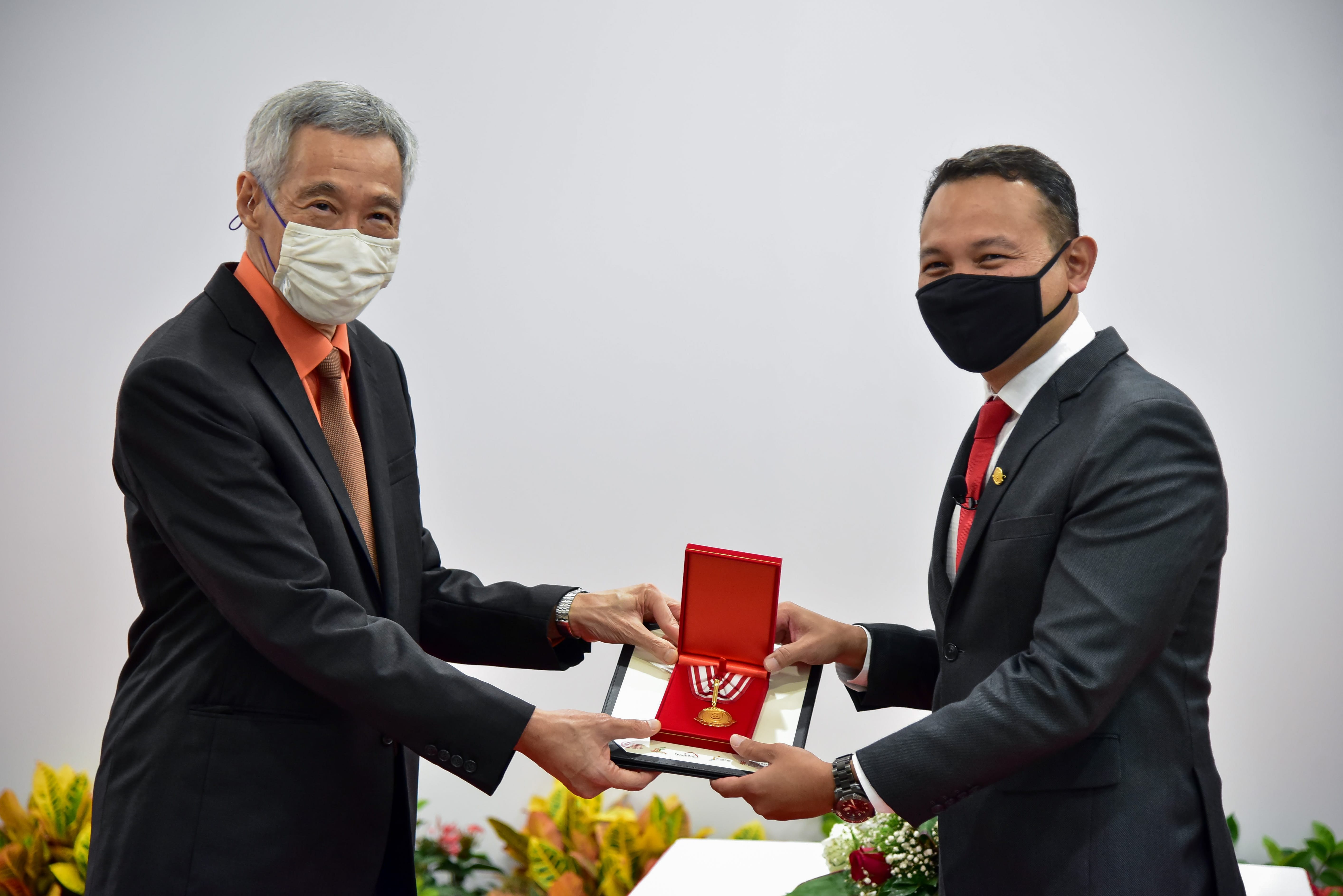 Two men in suits are holding a red award box containing a medal, and greenery is visible in the background.