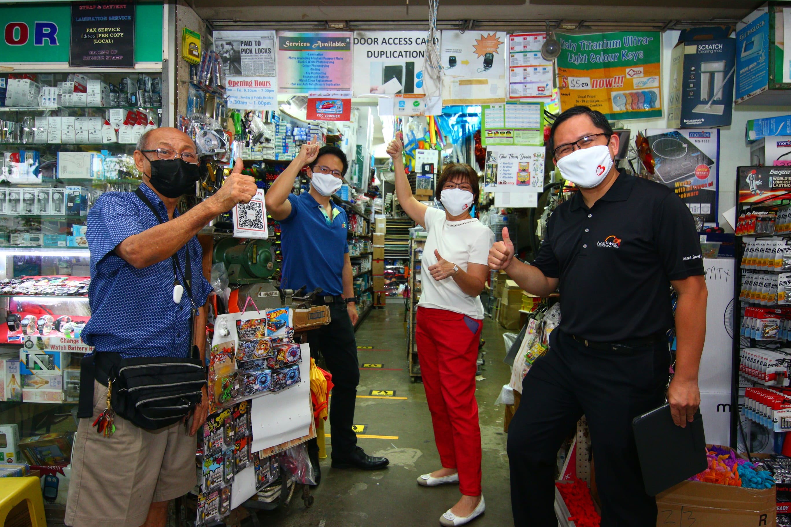 Four people inside a crowded shop are giving thumbs up; the store is filled with various merchandise and signs.