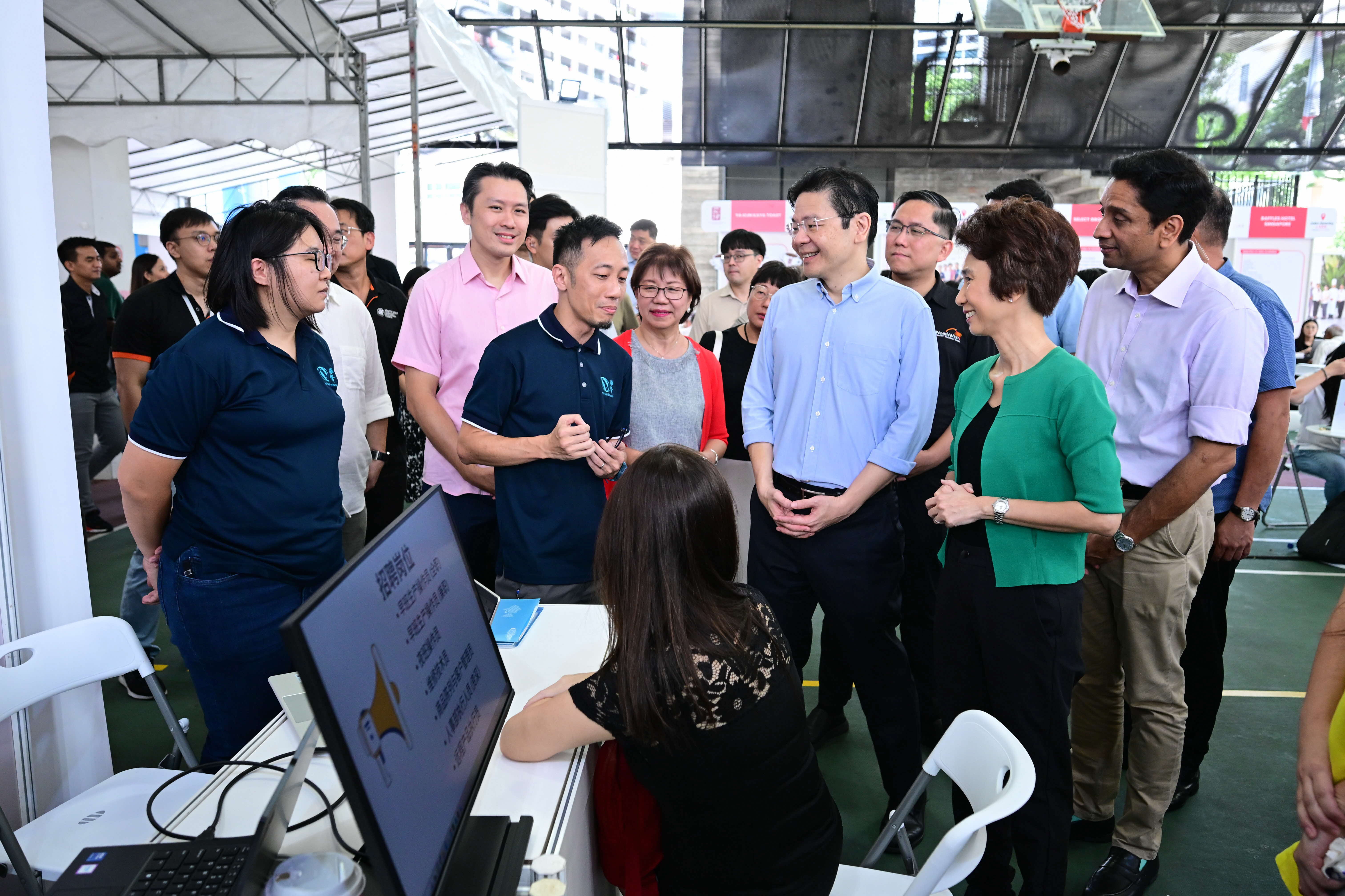 People standing and talking around a table with a seated individual working on a computer.