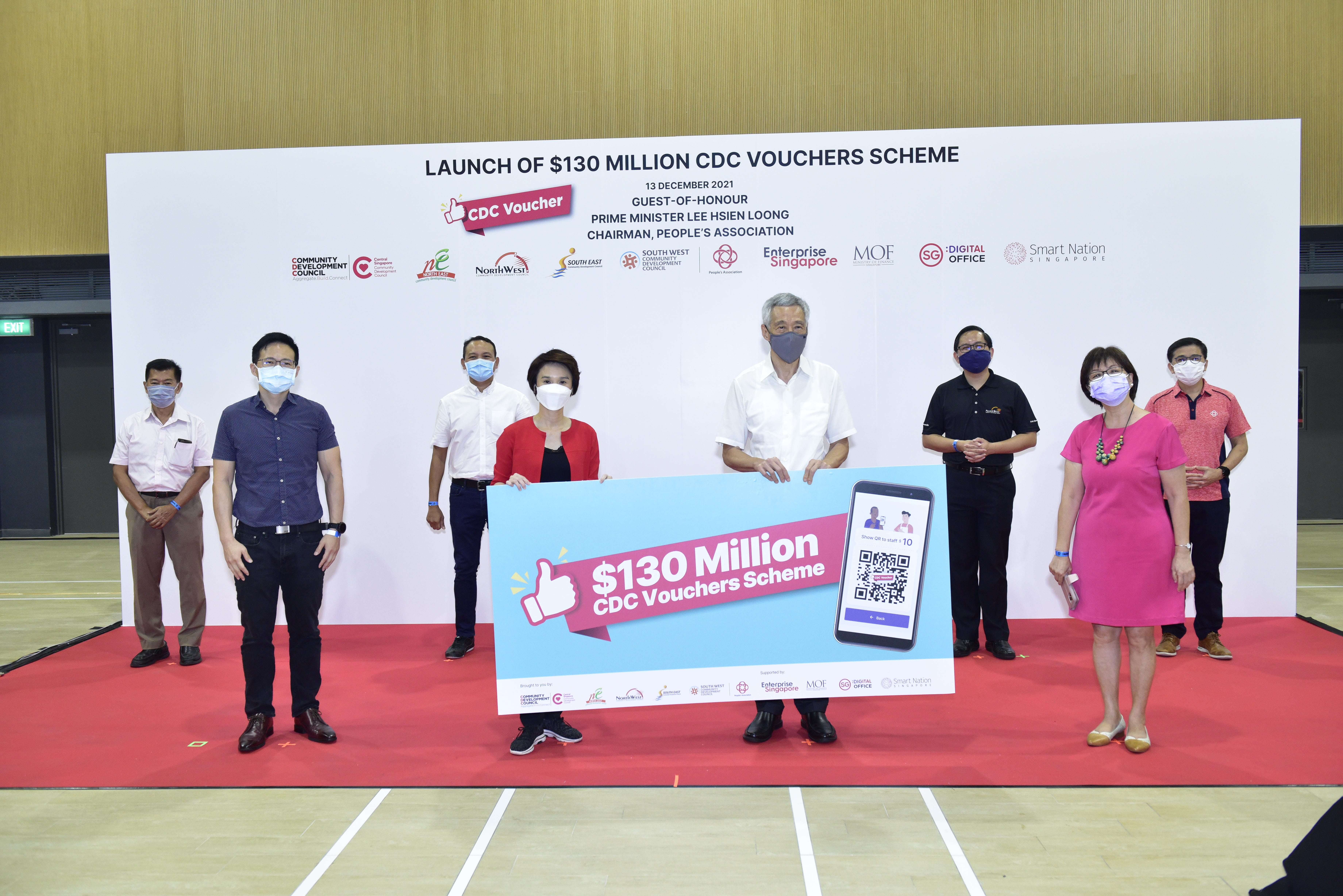 People stand on a red carpet holding a sign that reads "$130 Million CDC Vouchers Scheme" at an event.