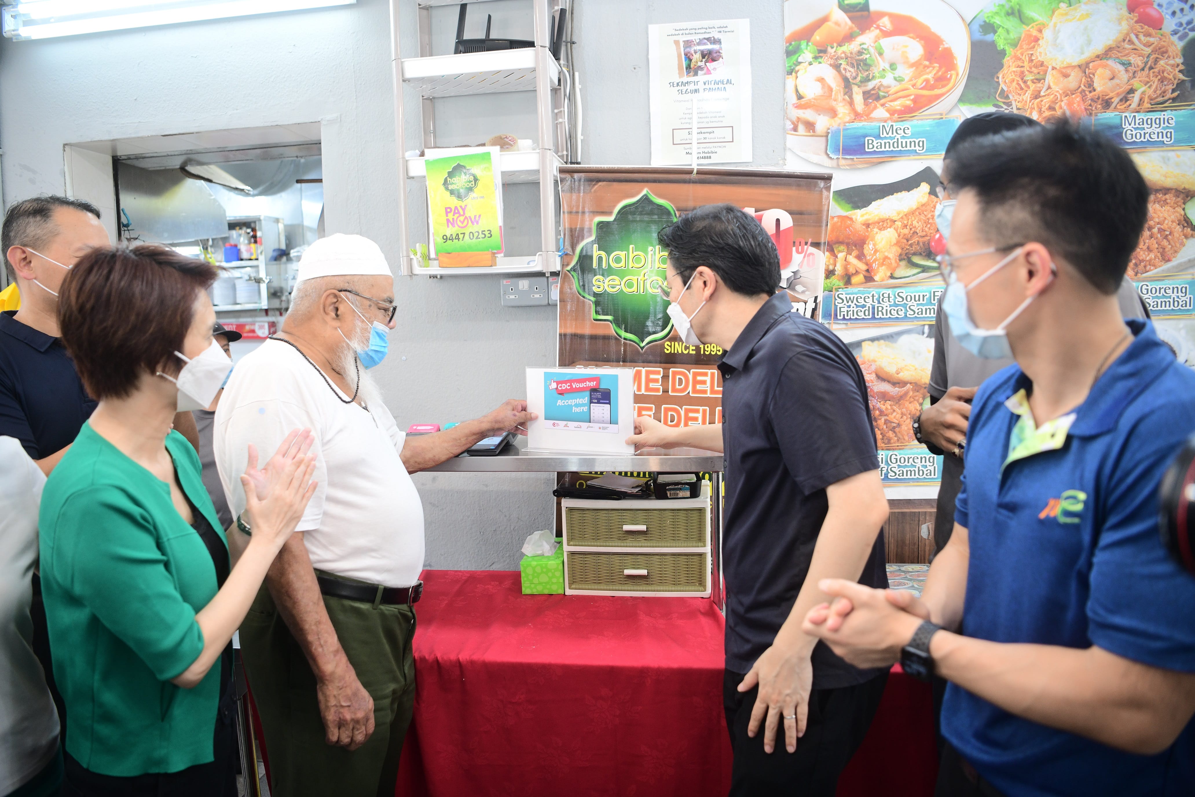 Several people watch an elderly man interacting with a digital voucher display in a food stall environment.