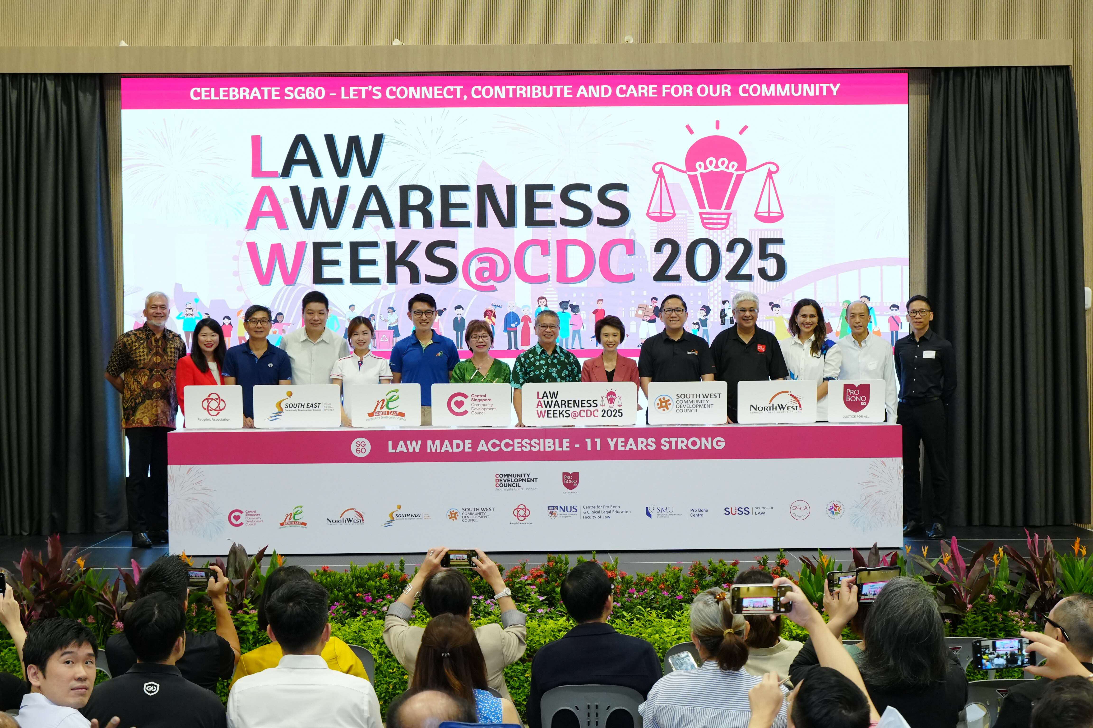 A group of people stand behind a table with various logos at a Law Awareness Weeks event; audience members are taking photos.