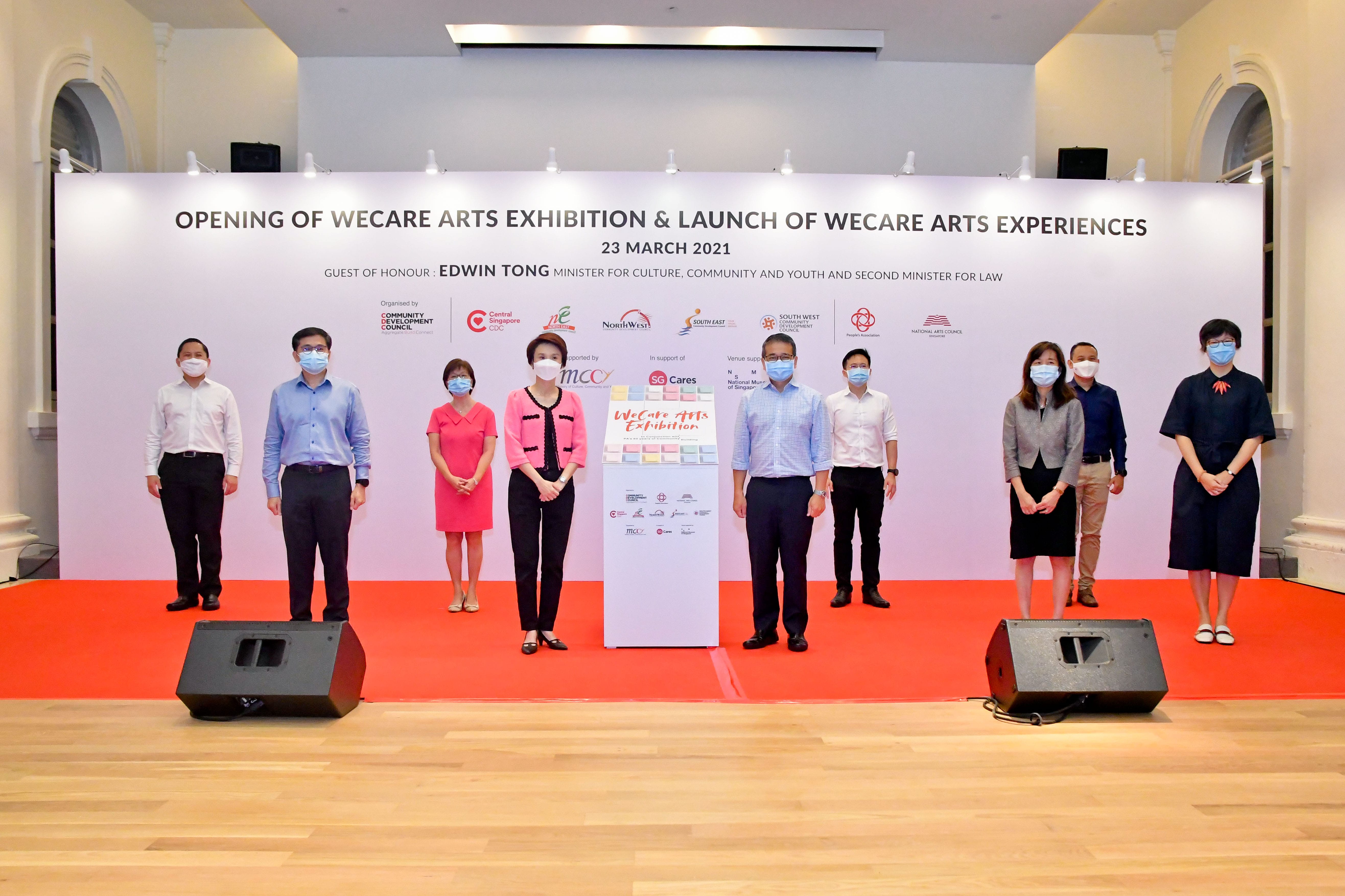 Nine people stand in front of a backdrop with text announcing the opening of an arts exhibition on March 23, 2021.