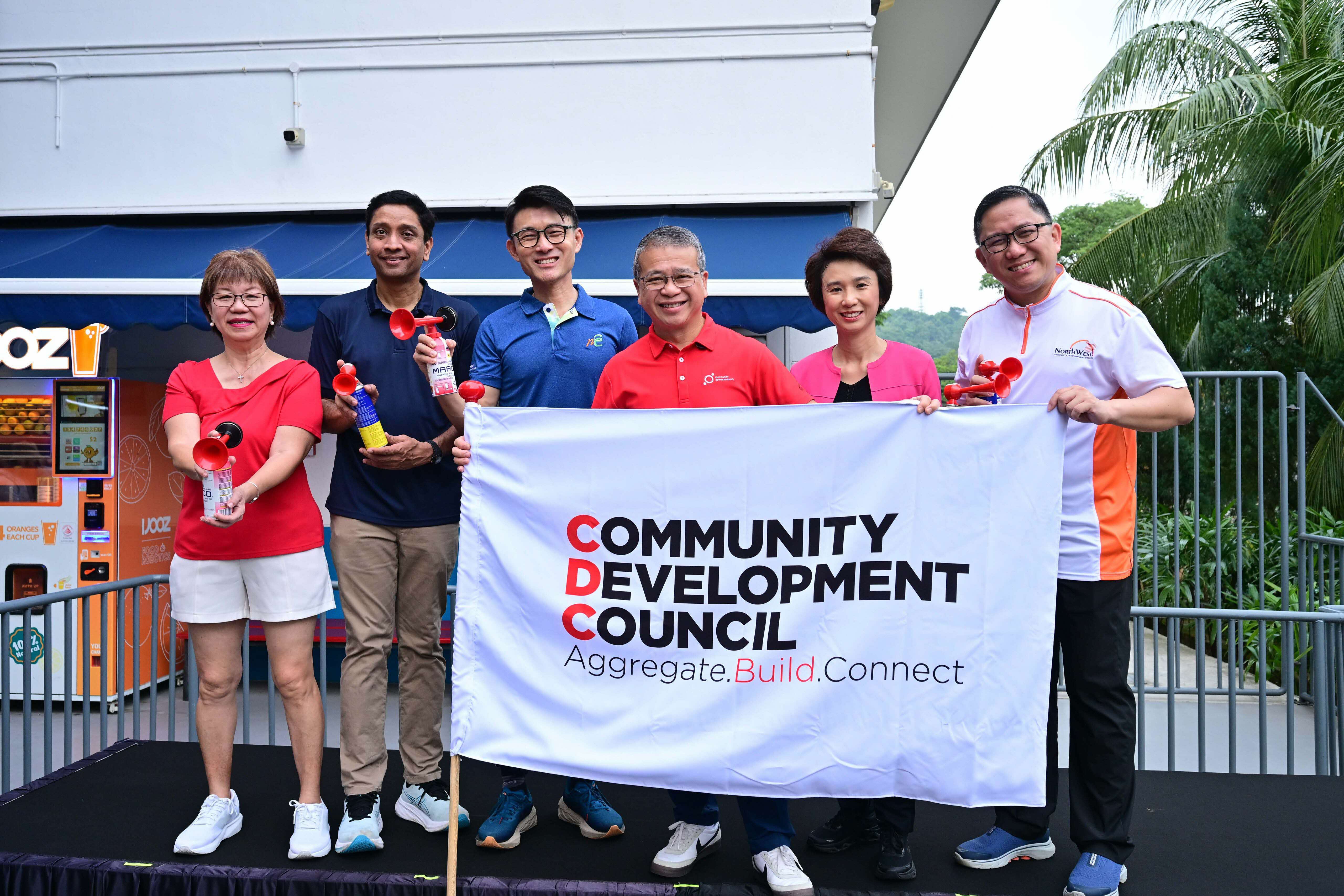 Seven people holding air horns stand behind a "Community Development Council" banner outdoors.