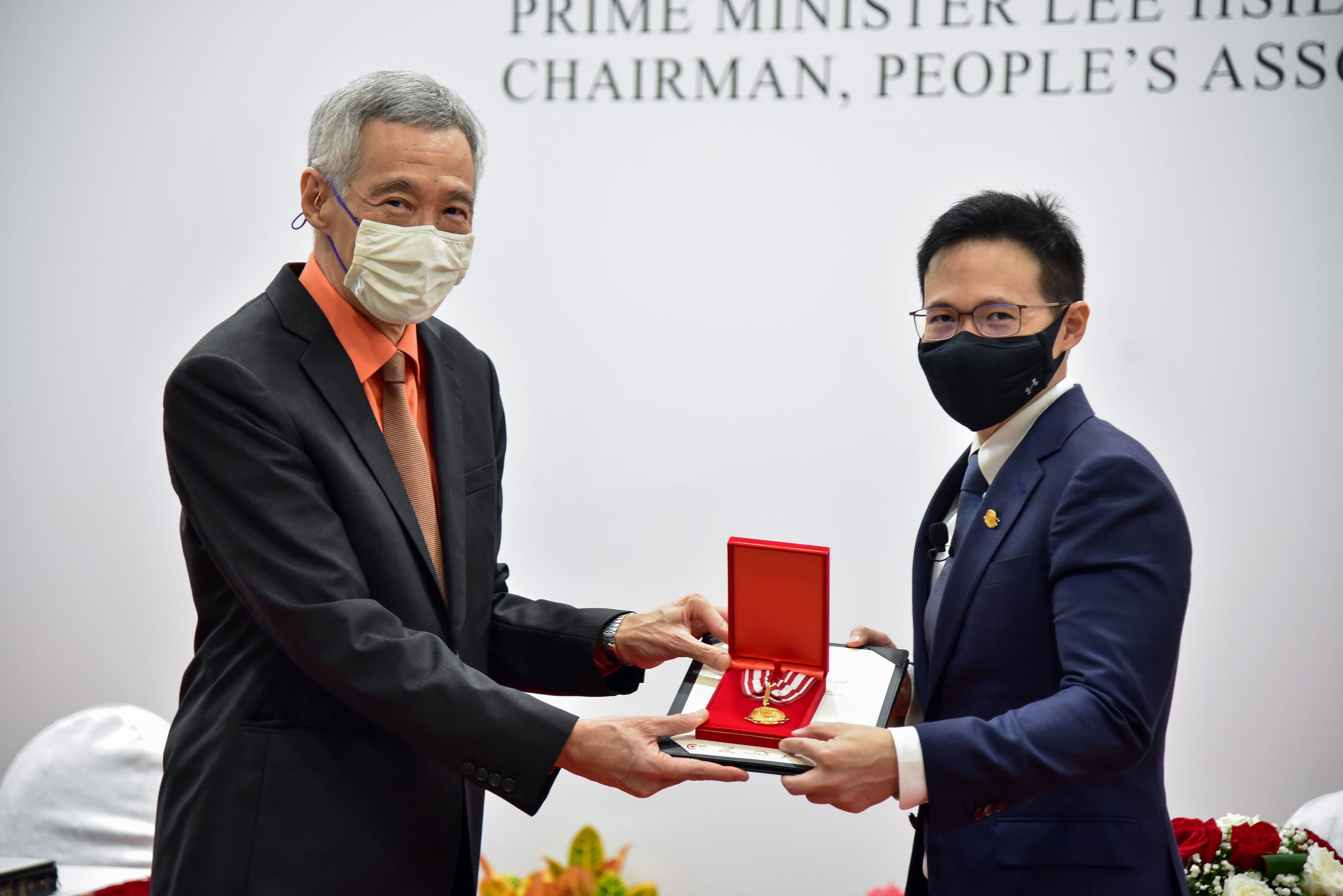 Two individuals exchange a red award box while standing in front of a backdrop with text.