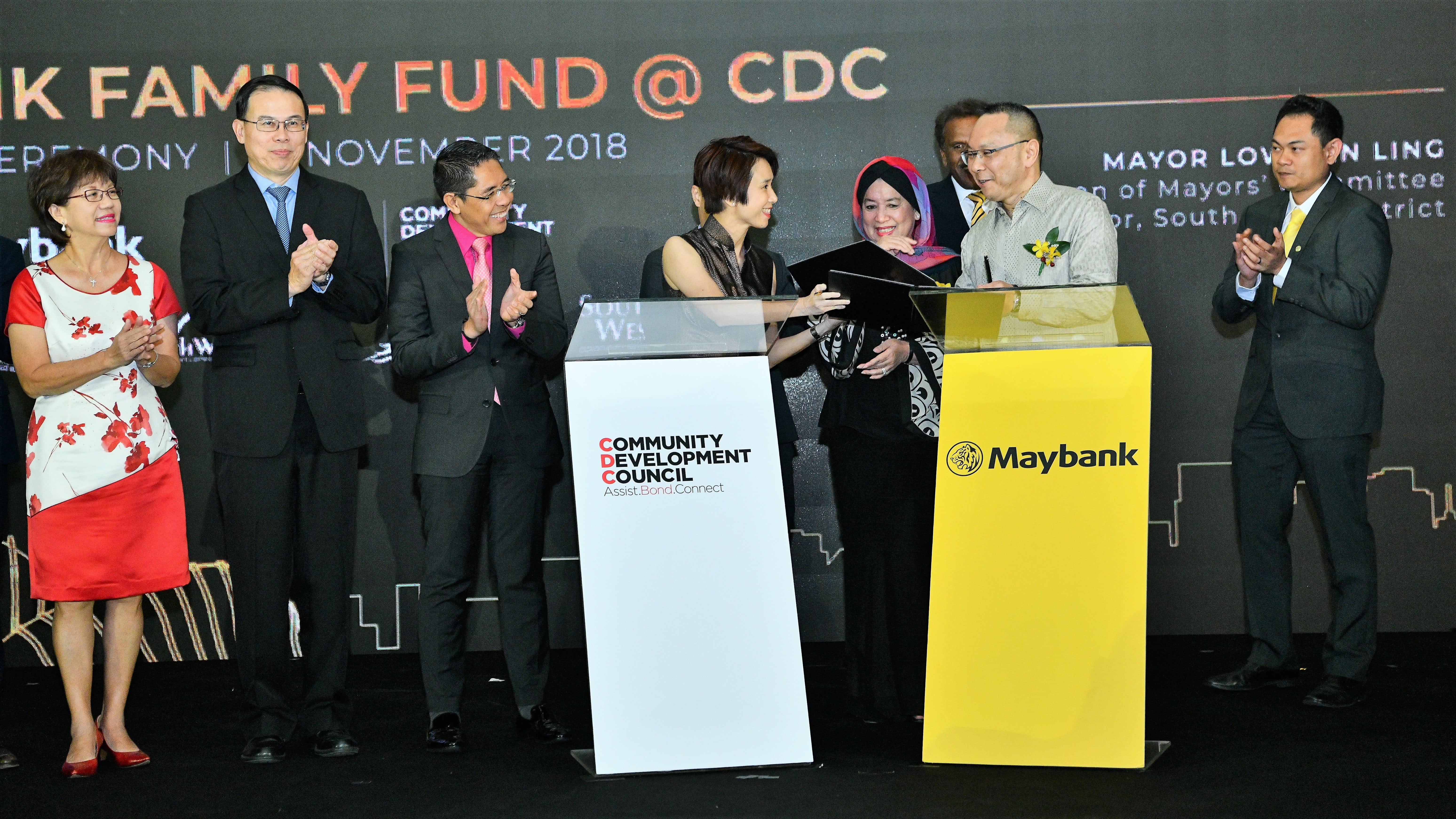 People are standing and clapping at an event with signs for Community Development Council and Maybank.