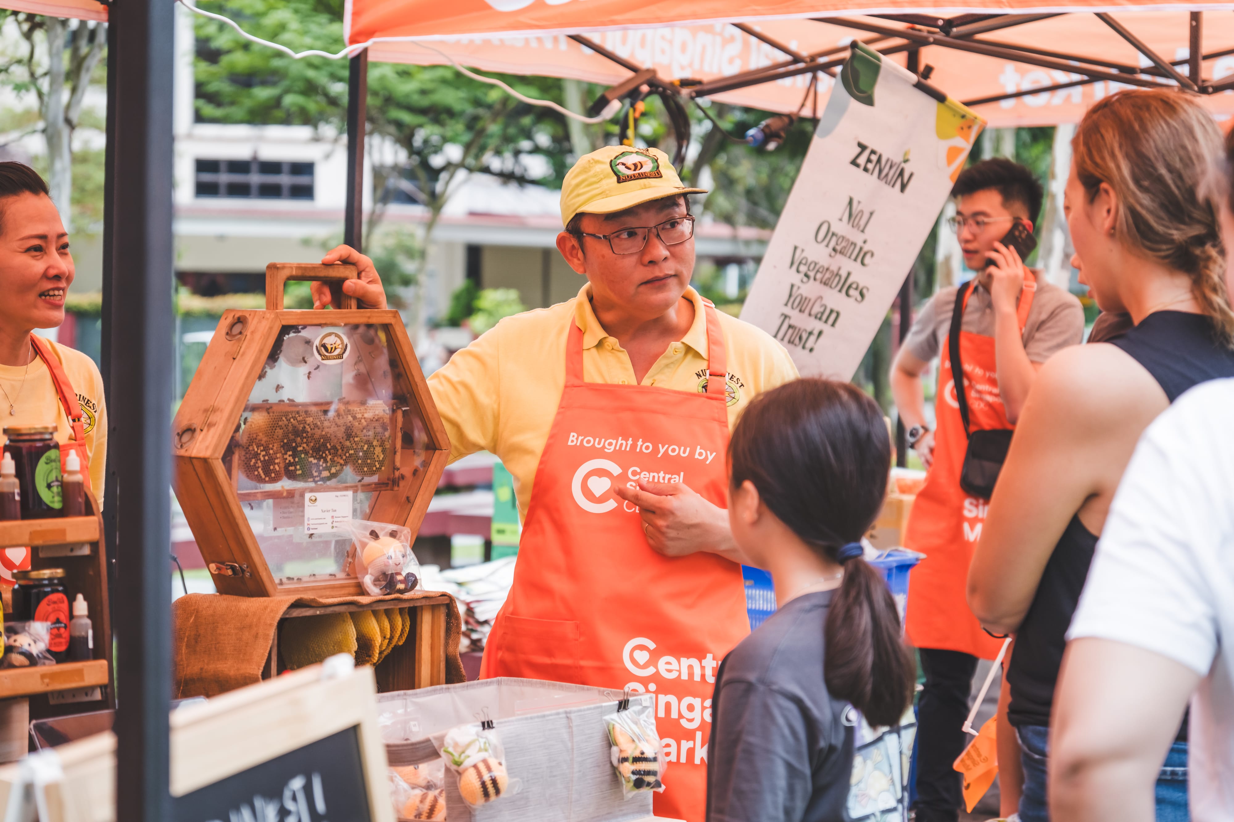 A market vendor shows honeycomb in a wooden frame filled with live bees, surrounded by customers and products under orange tents.