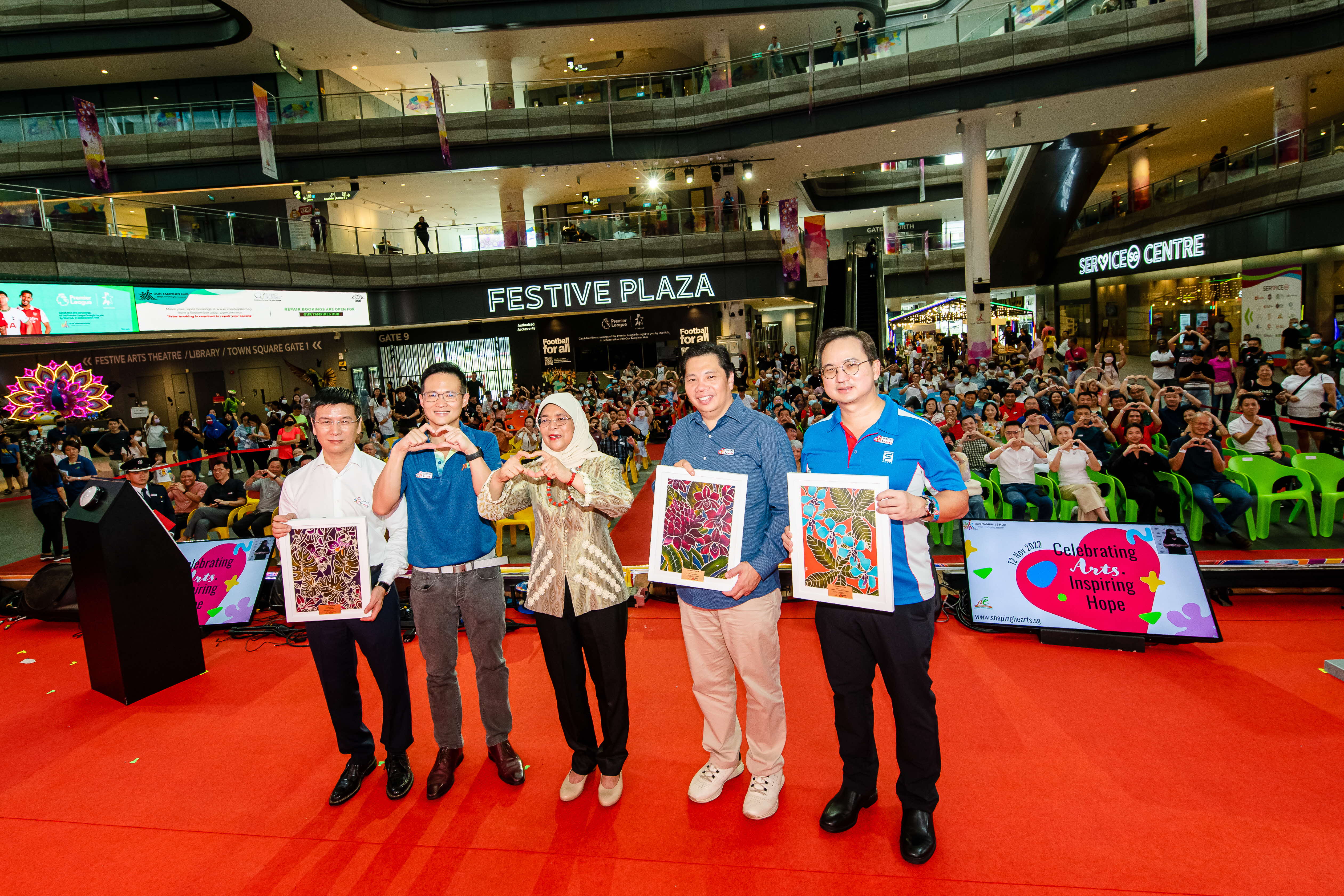 Five people stand on a red stage holding artwork, with a large crowd seated behind them in Festive Plaza.