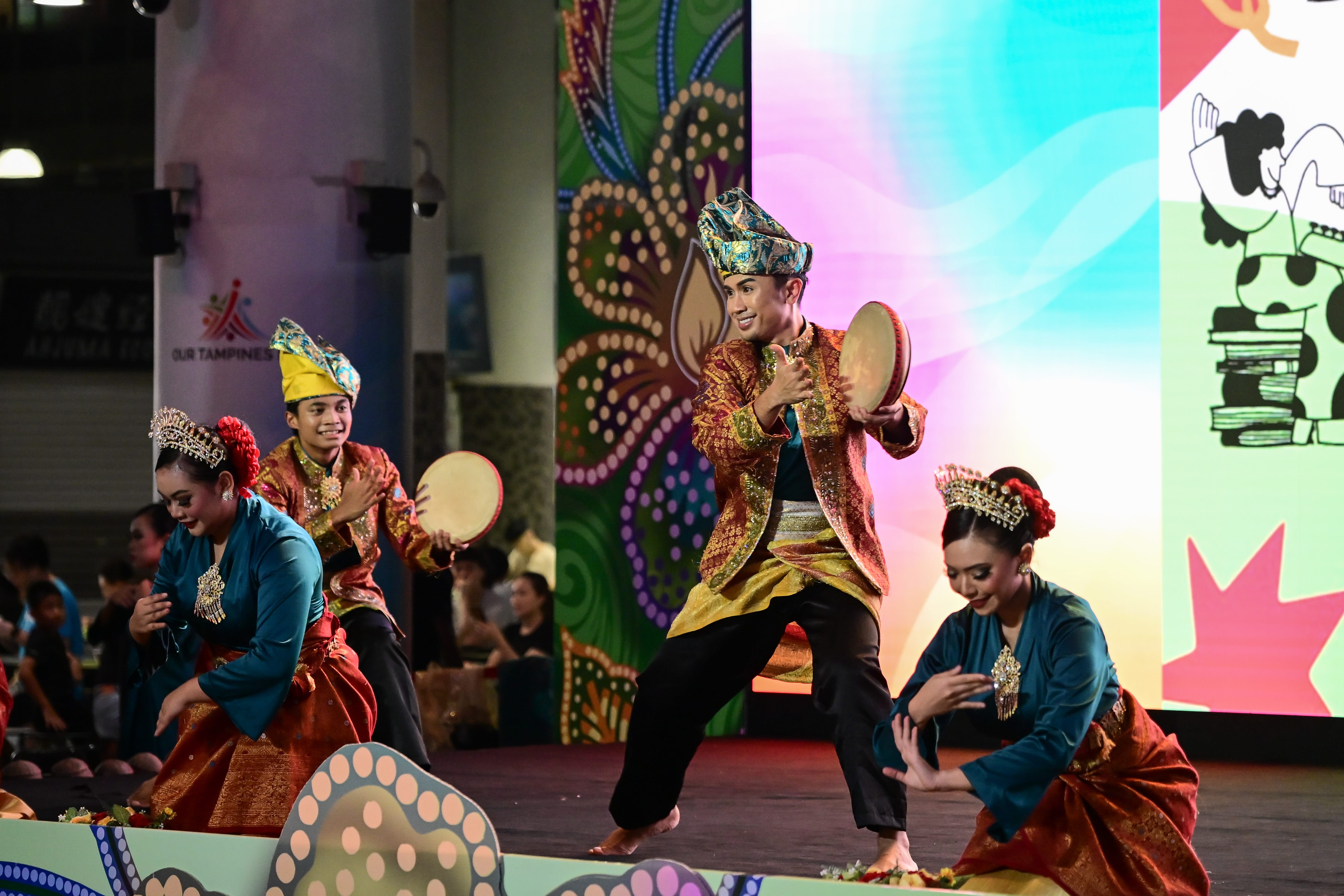Performers in colorful traditional clothing and headpieces with drums, on stage, against a vibrant, patterned backdrop.