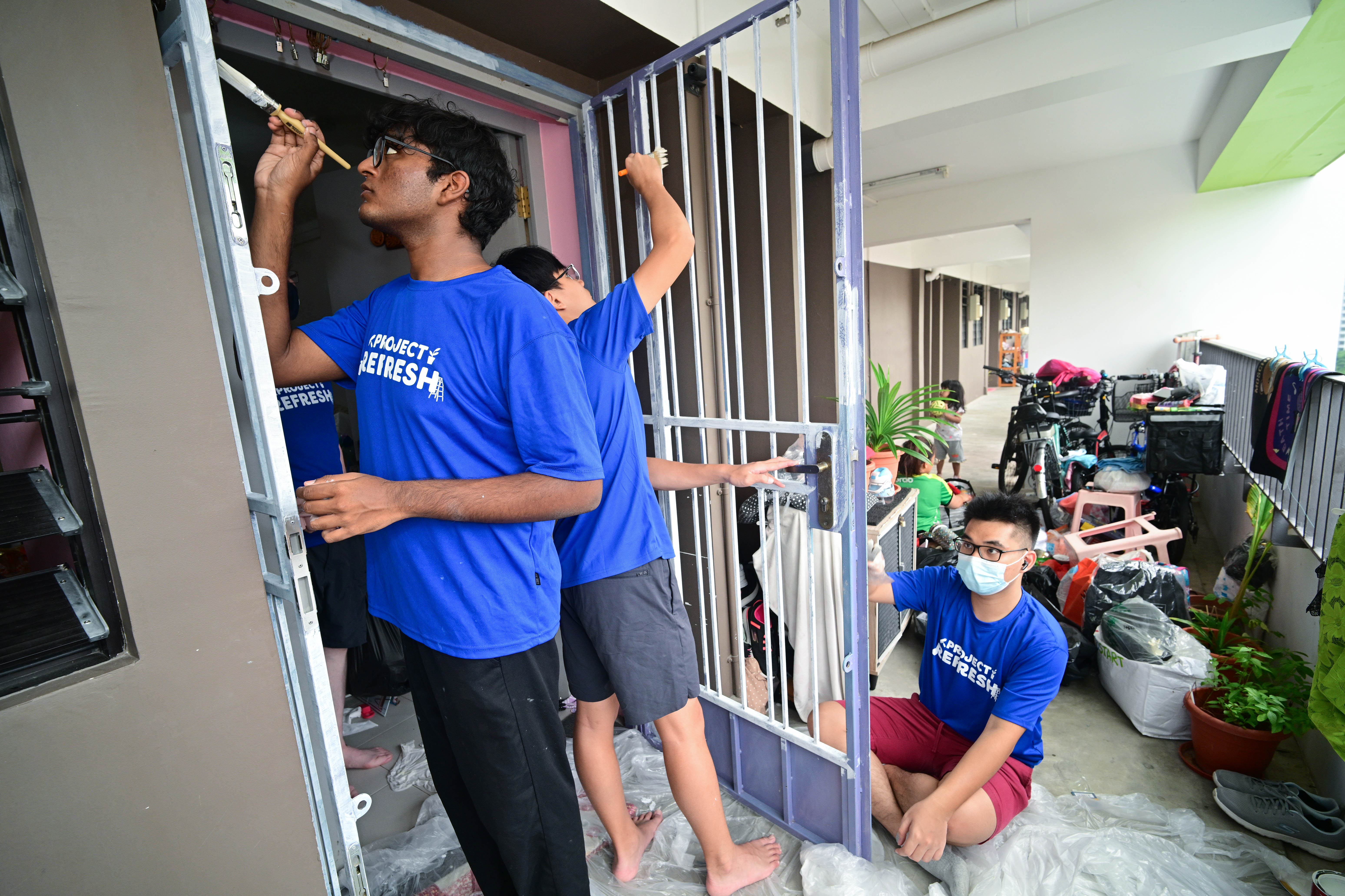 Several people in blue shirts are painting a metal gate in a hallway filled with various items.