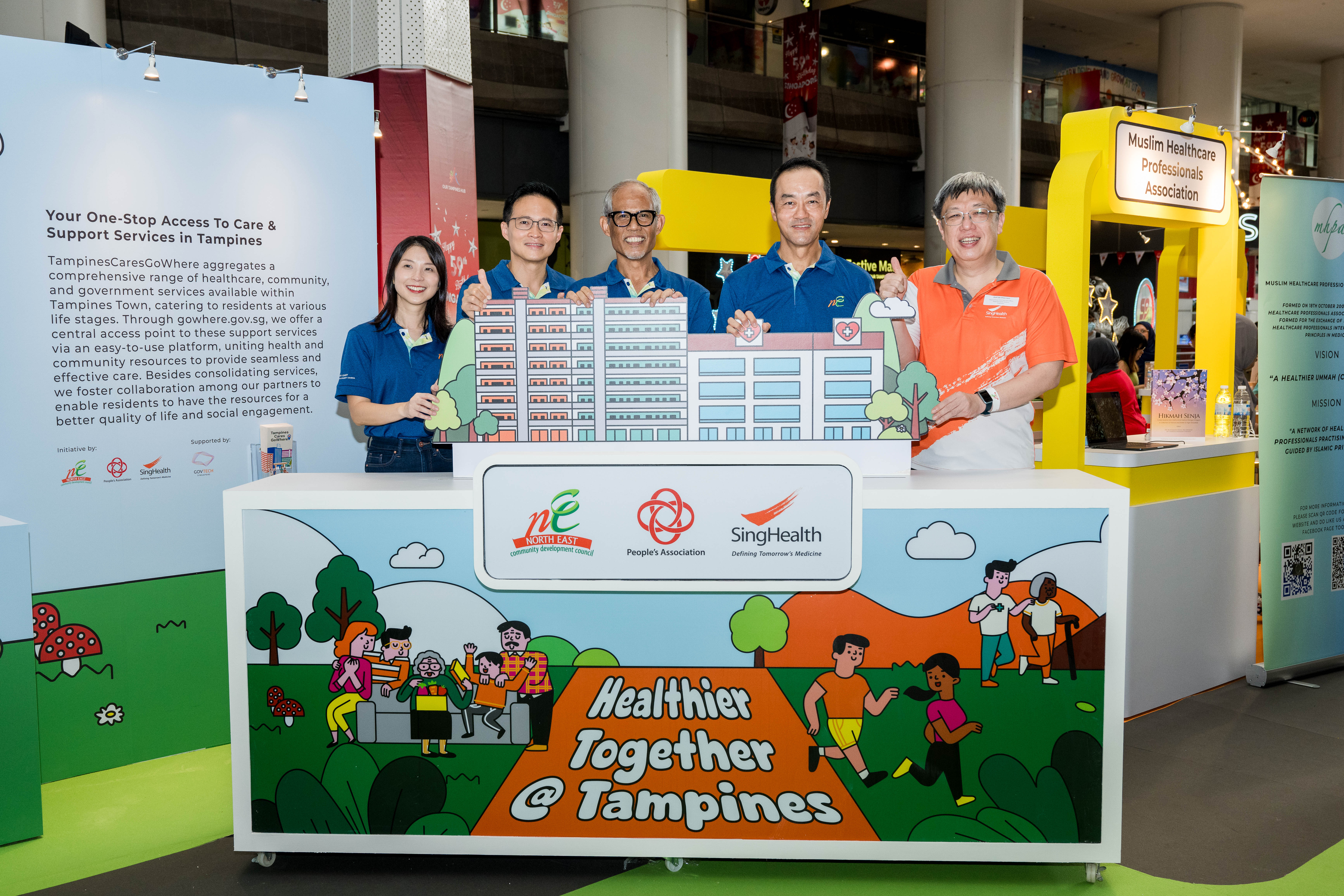 Six people stand behind a display table with illustrations and health-related logos in a public indoor space.