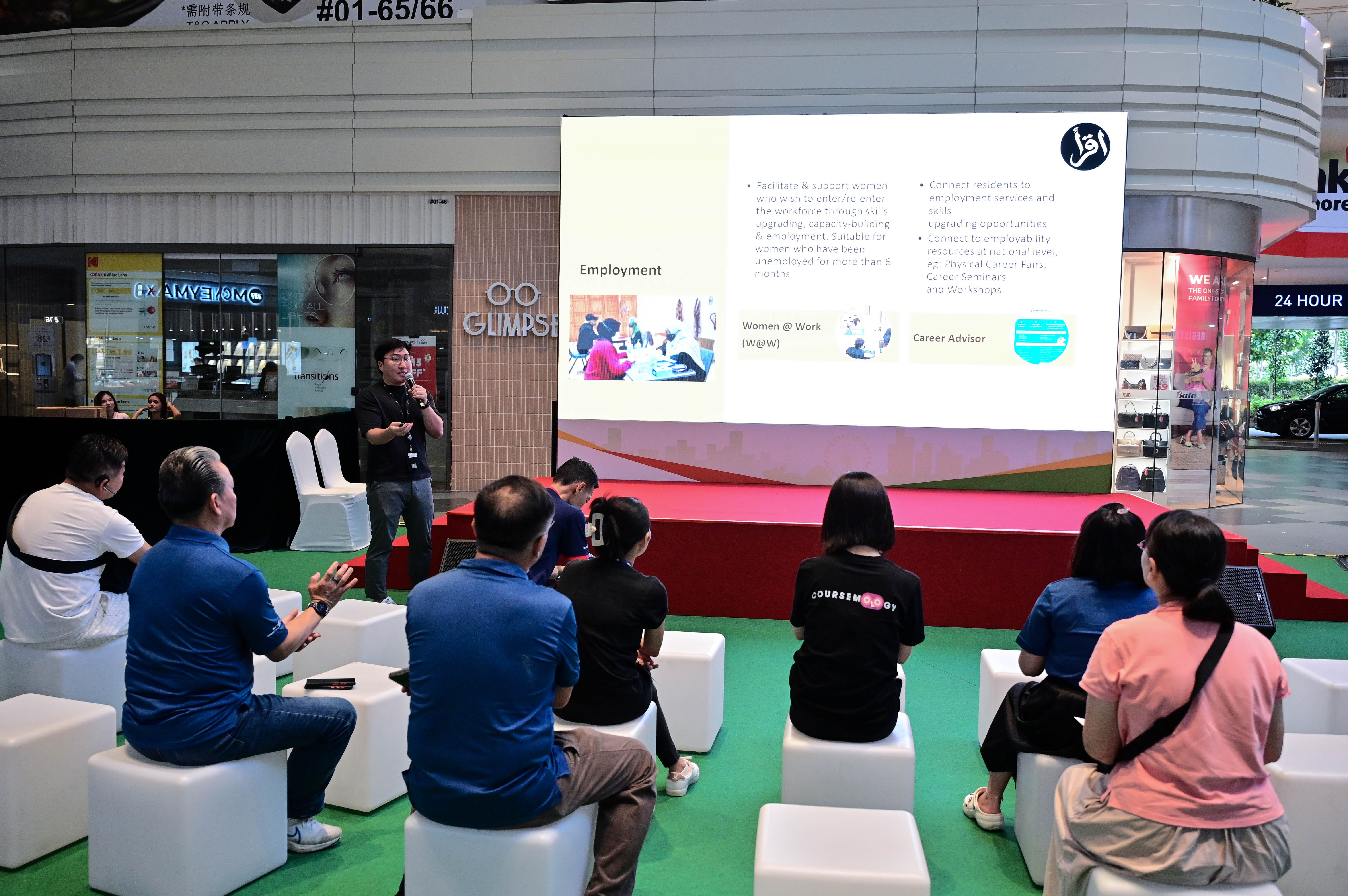 A group of people is watching a person presenting information about employment and career advice on a large screen in a mall.
