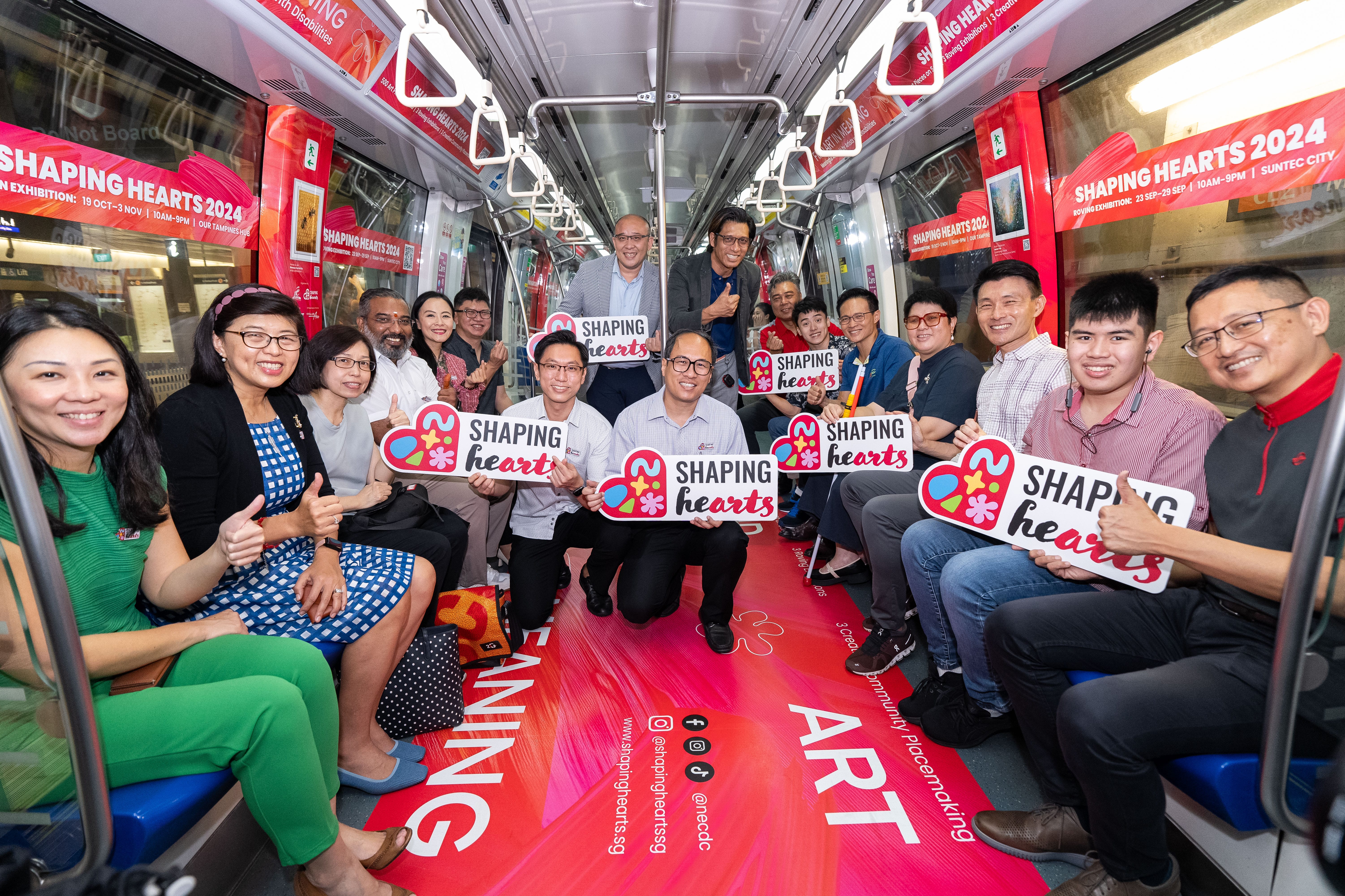People on a train holding "Shaping Hearts" signs with red advertisements for "Shaping Hearts 2024" in the background.