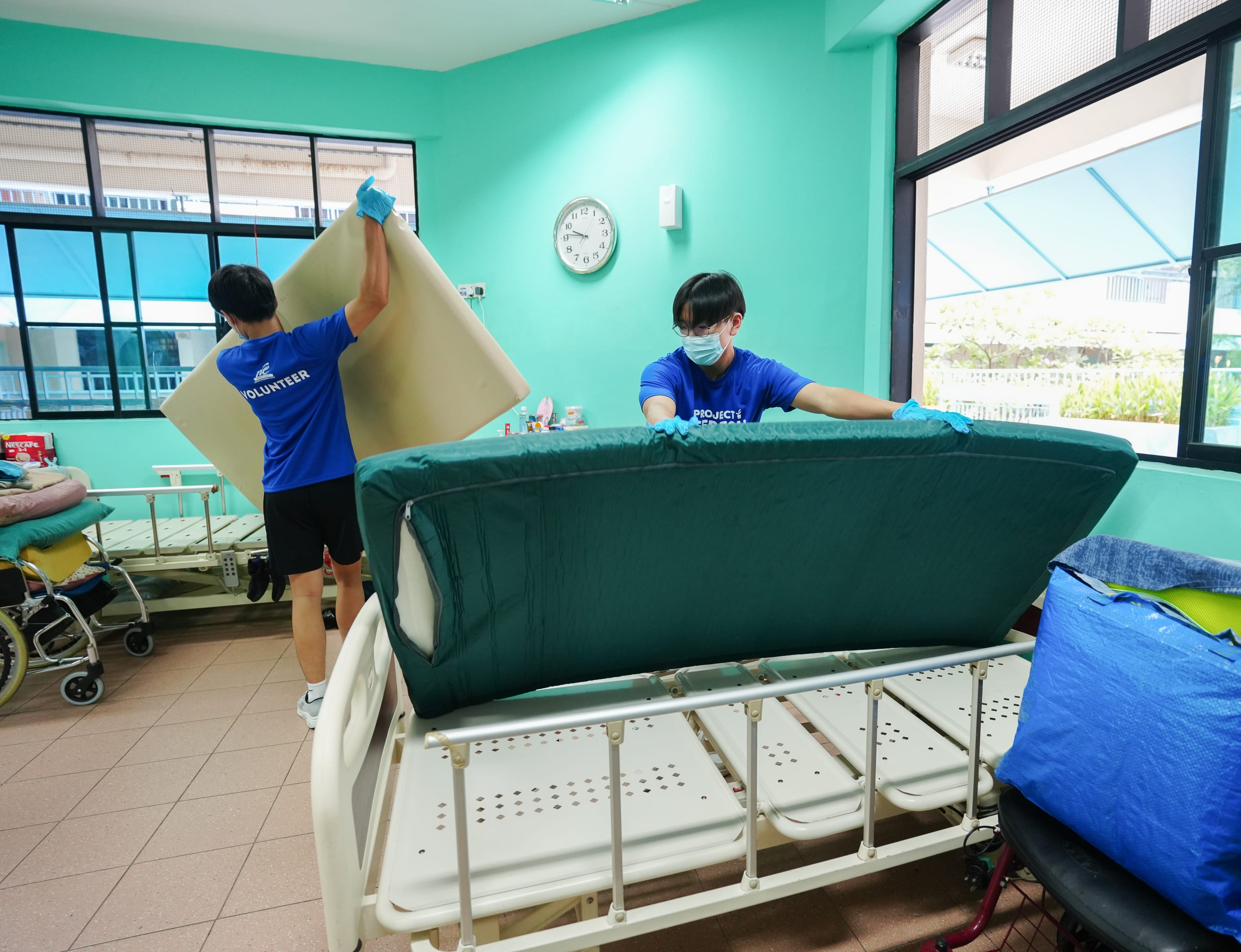 Two people in blue shirts are lifting a mattress in a room with green walls and large windows.
