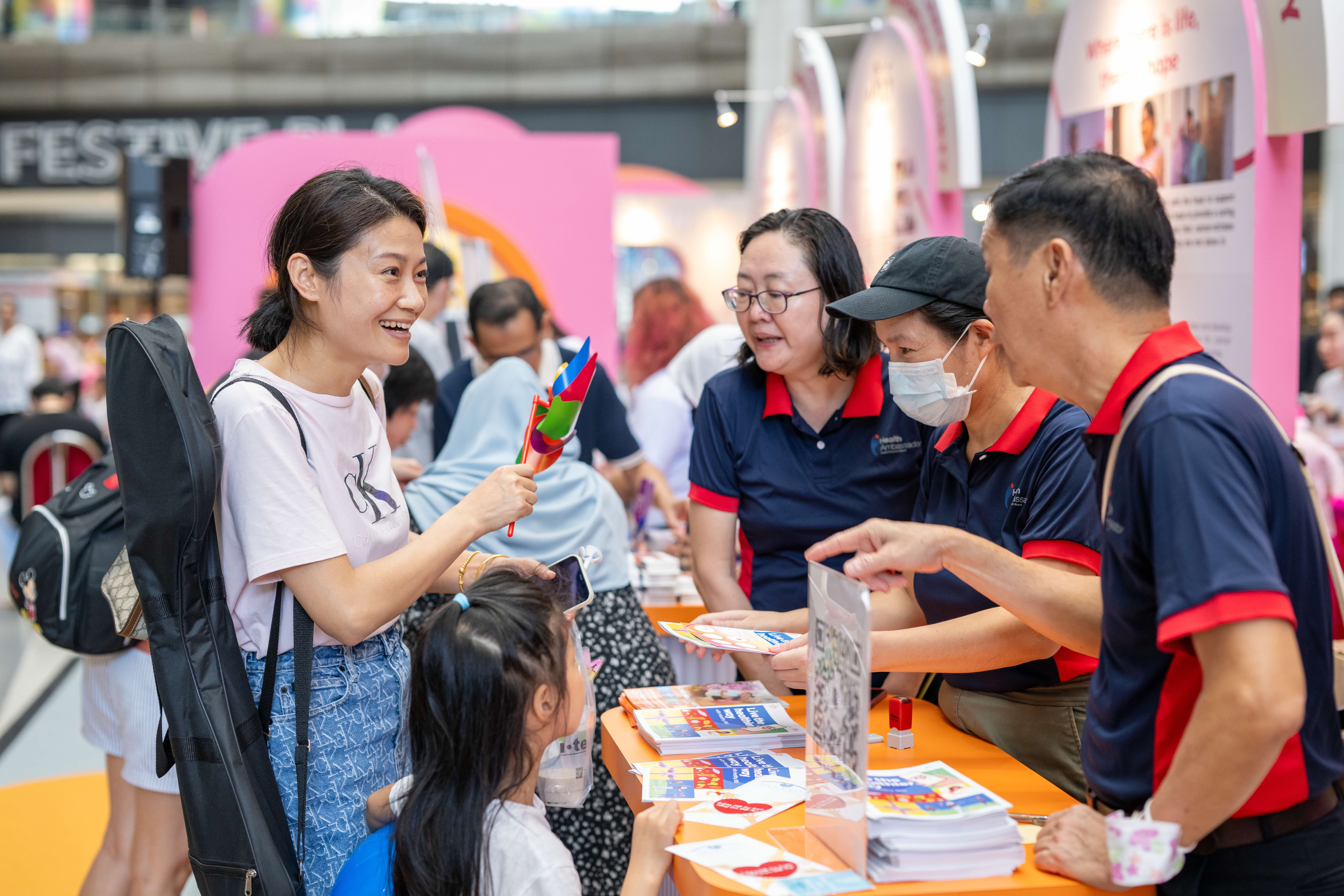 A woman holding a pinwheel talks to three people wearing navy shirts with red collars at a booth, while a child looks on.
