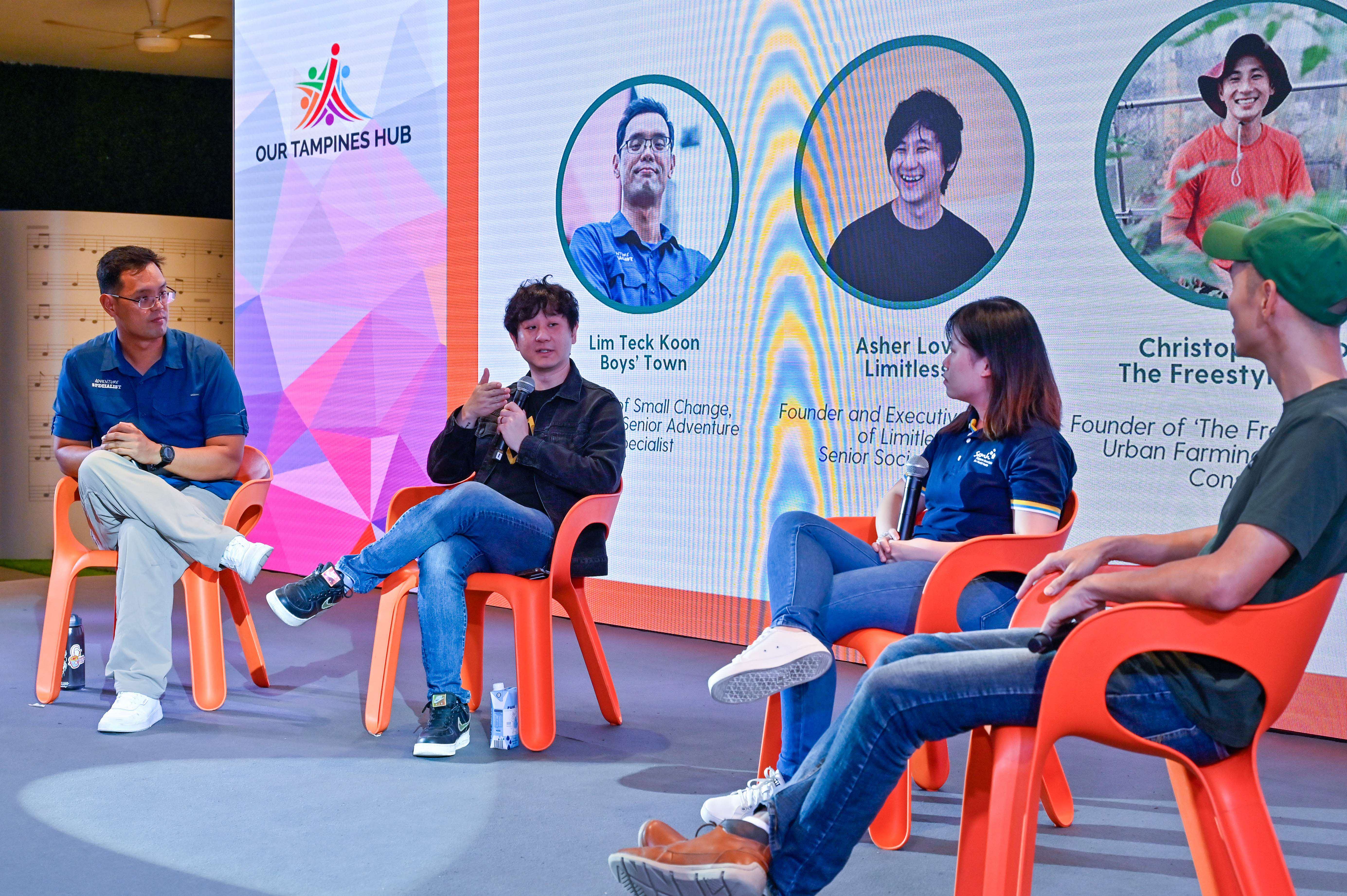 Four people sit on orange chairs on a stage with a large screen displaying text and graphics behind them.