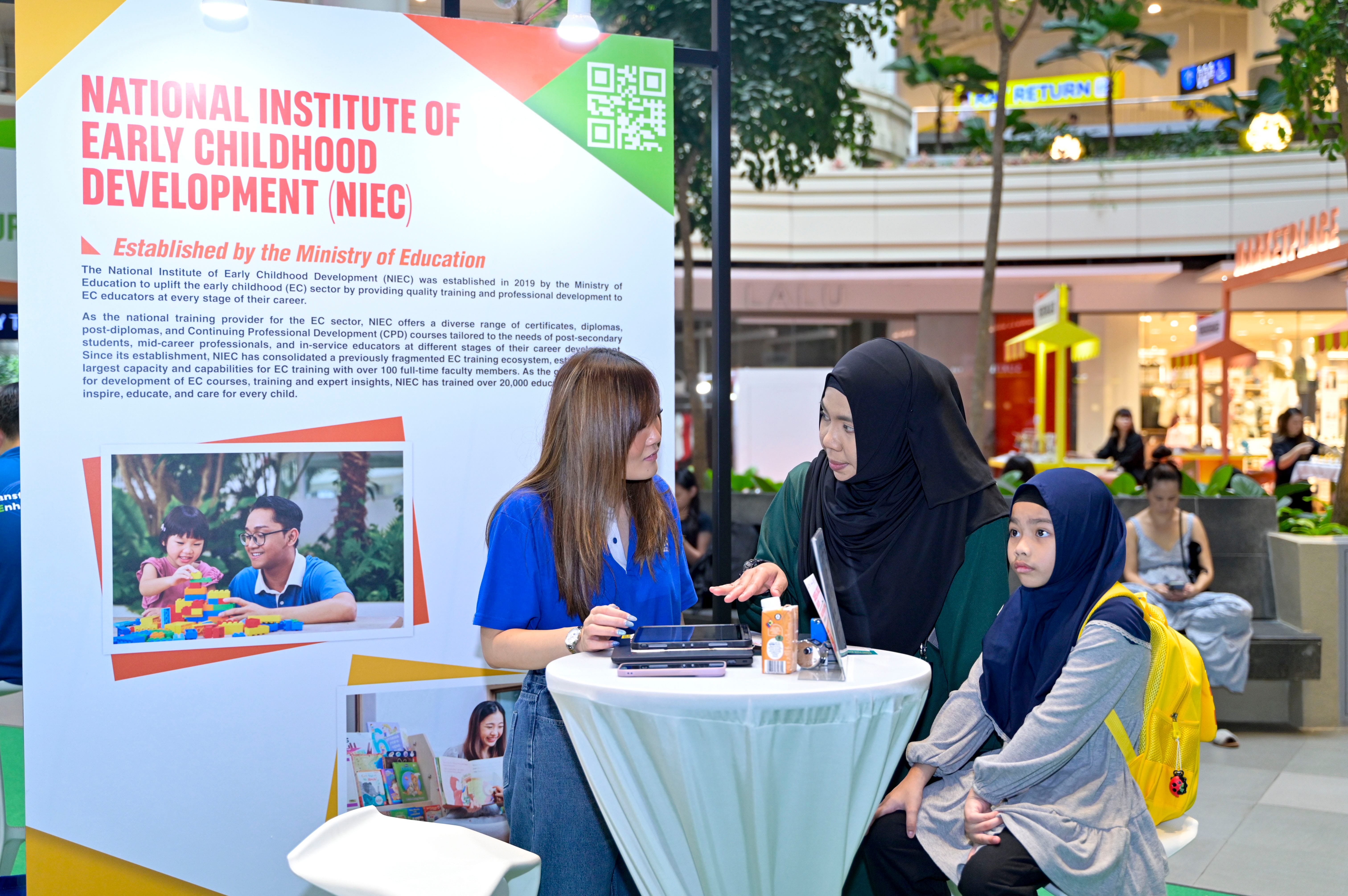 Two women and a child sit at a table in front of a National Institute of Early Childhood Development (NIEC) poster.