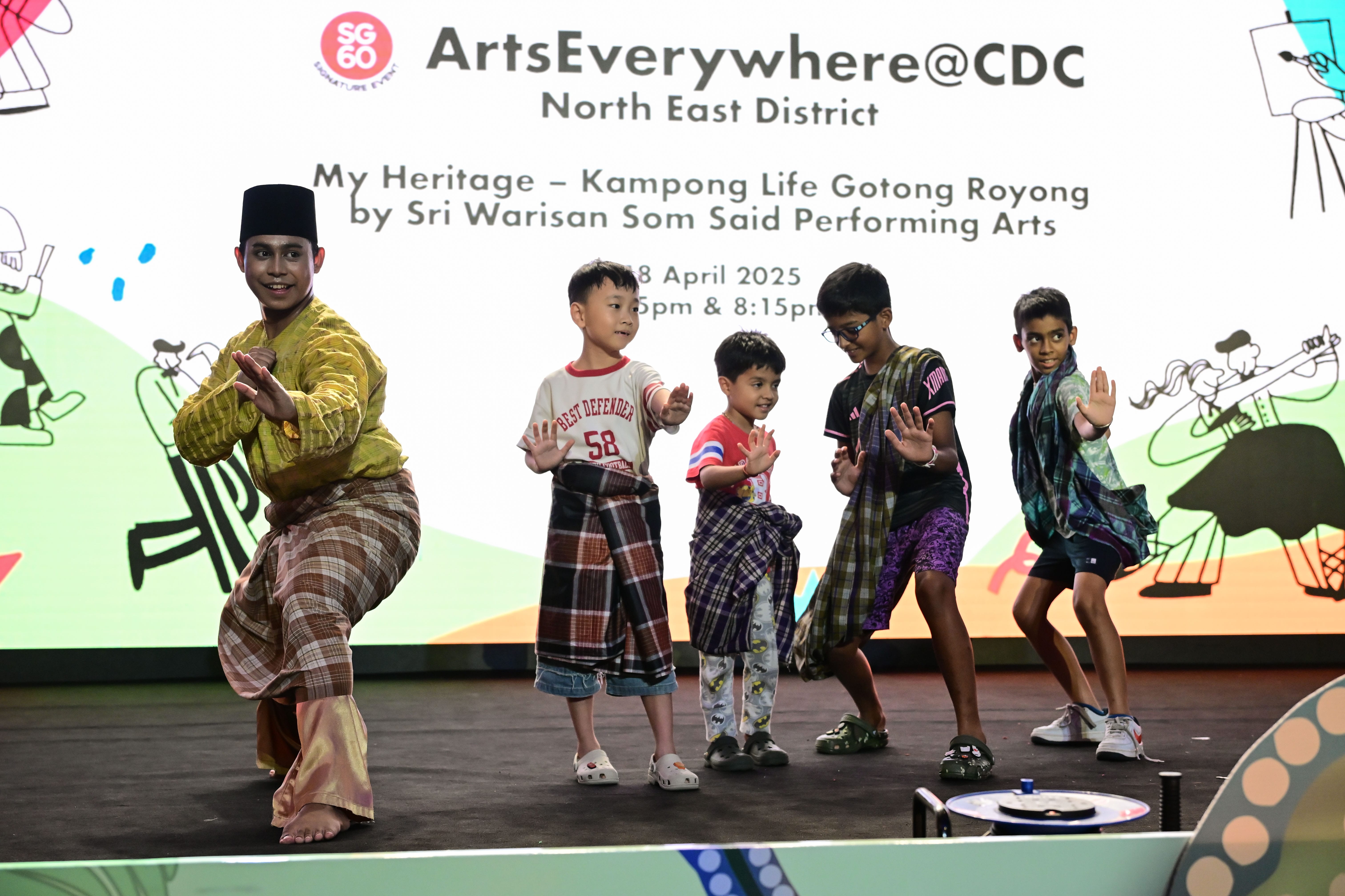 Ae performer in traditional attire strike poses while children beside him attempt to mimic him on stage with backgrounds promoting ArtsEverywhere@CDC event.
