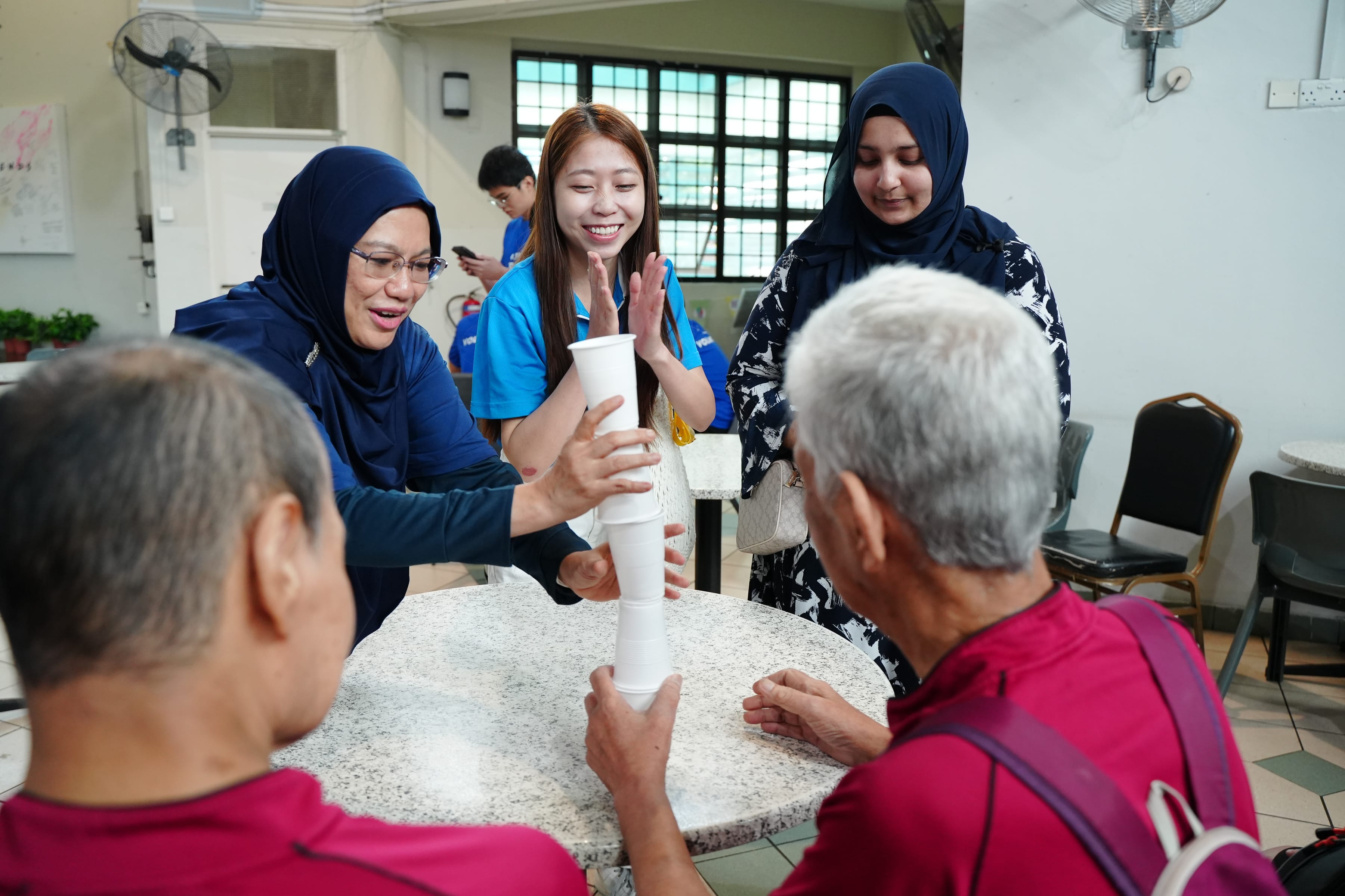 Several people stand around a table, stacking white cups in a line vertically.
