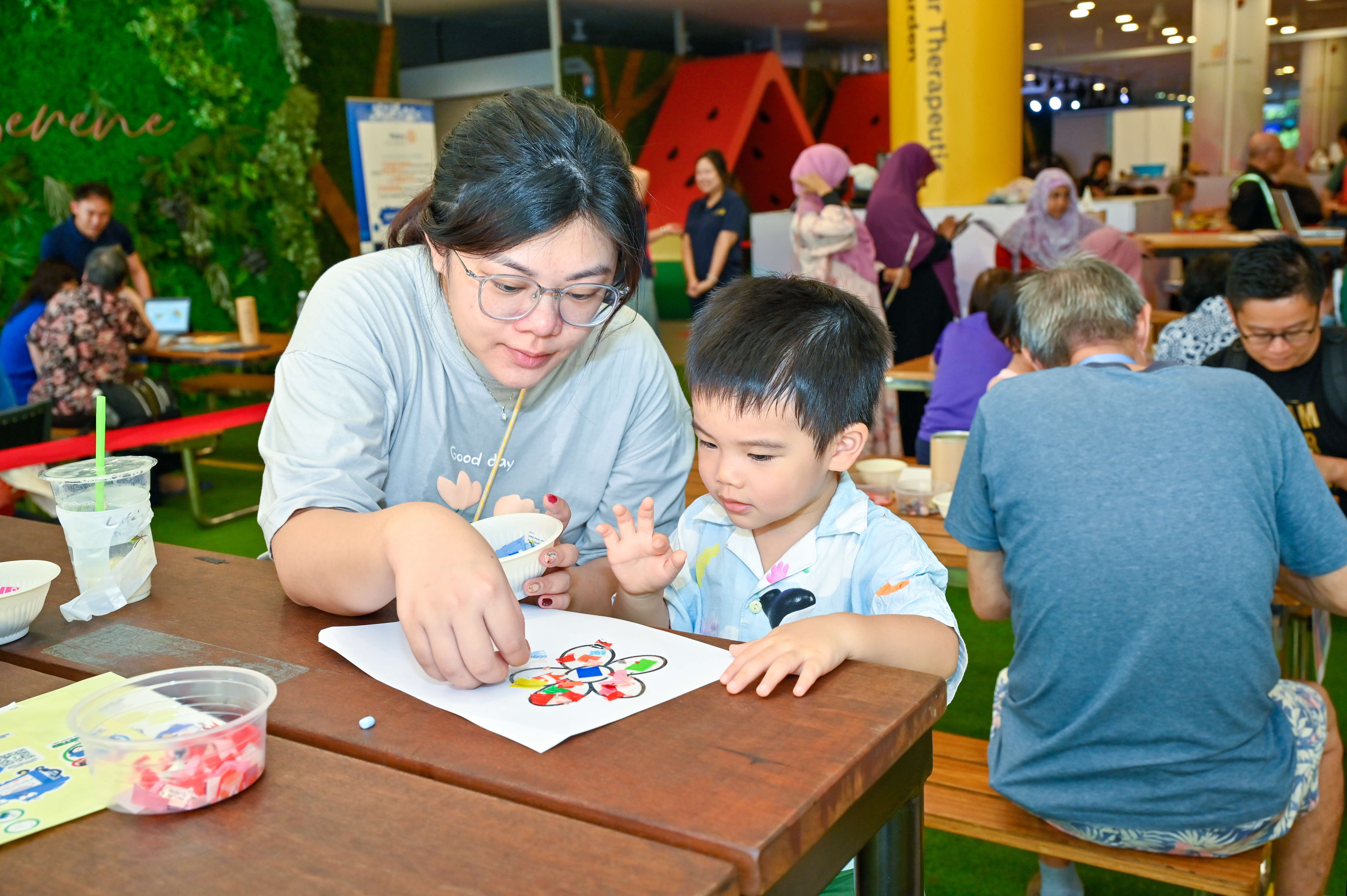 A child and an adult are coloring together at a table, surrounded by other people in a busy indoor setting.