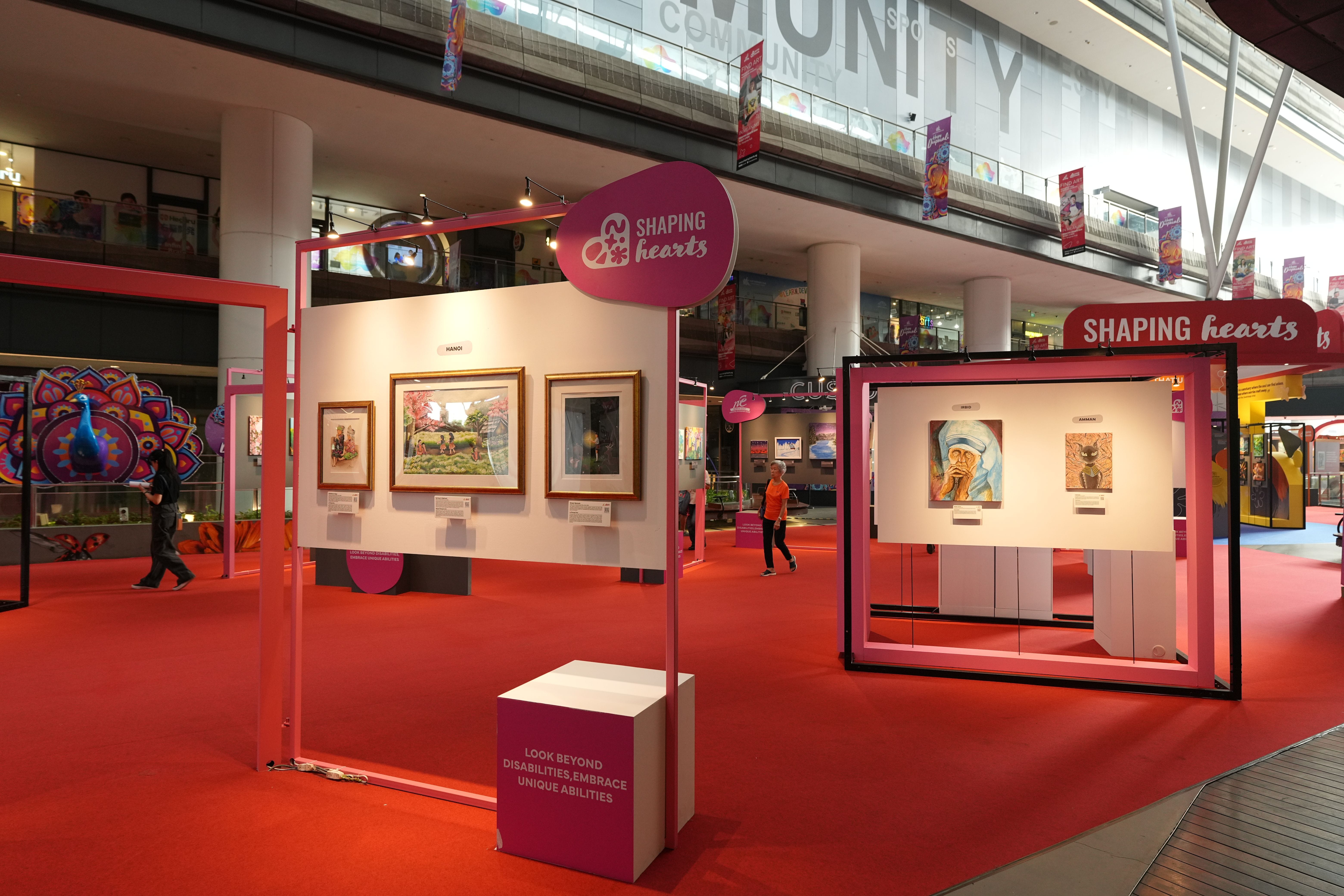 An art exhibition with framed paintings displayed in a brightly lit, spacious gallery with red flooring and banners.