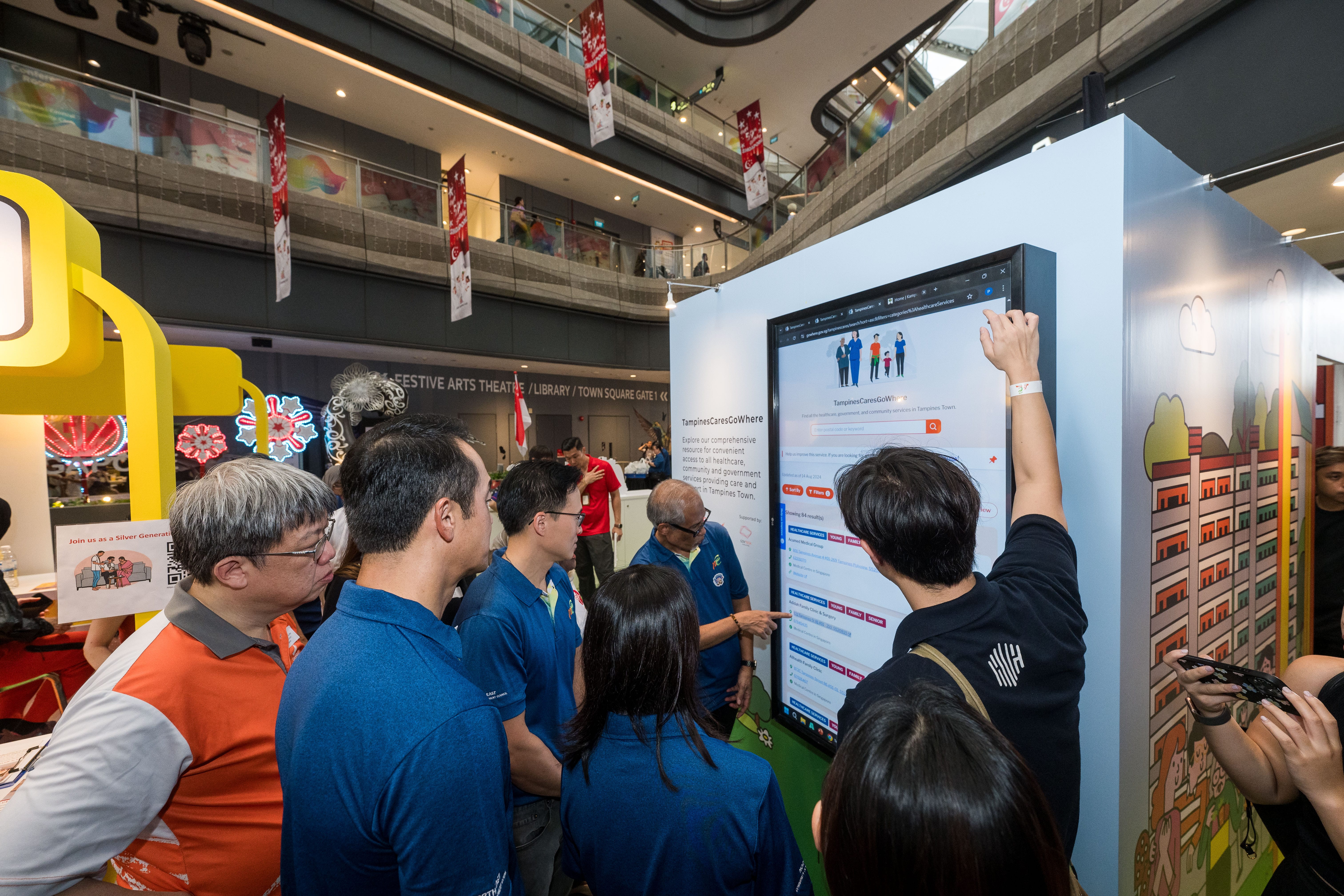 A group of people examine a large interactive touchscreen display in a spacious, multi-level indoor venue.