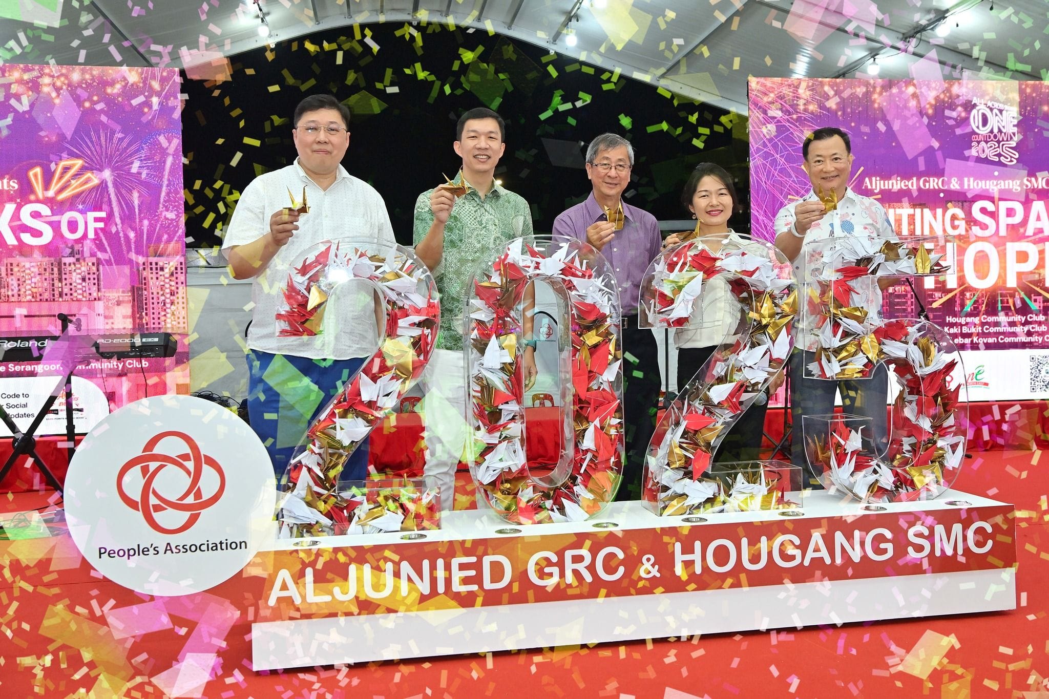 Five people hold gold origami cranes, standing behind large "2023" figures filled with origami cranes amidst colorful confetti.