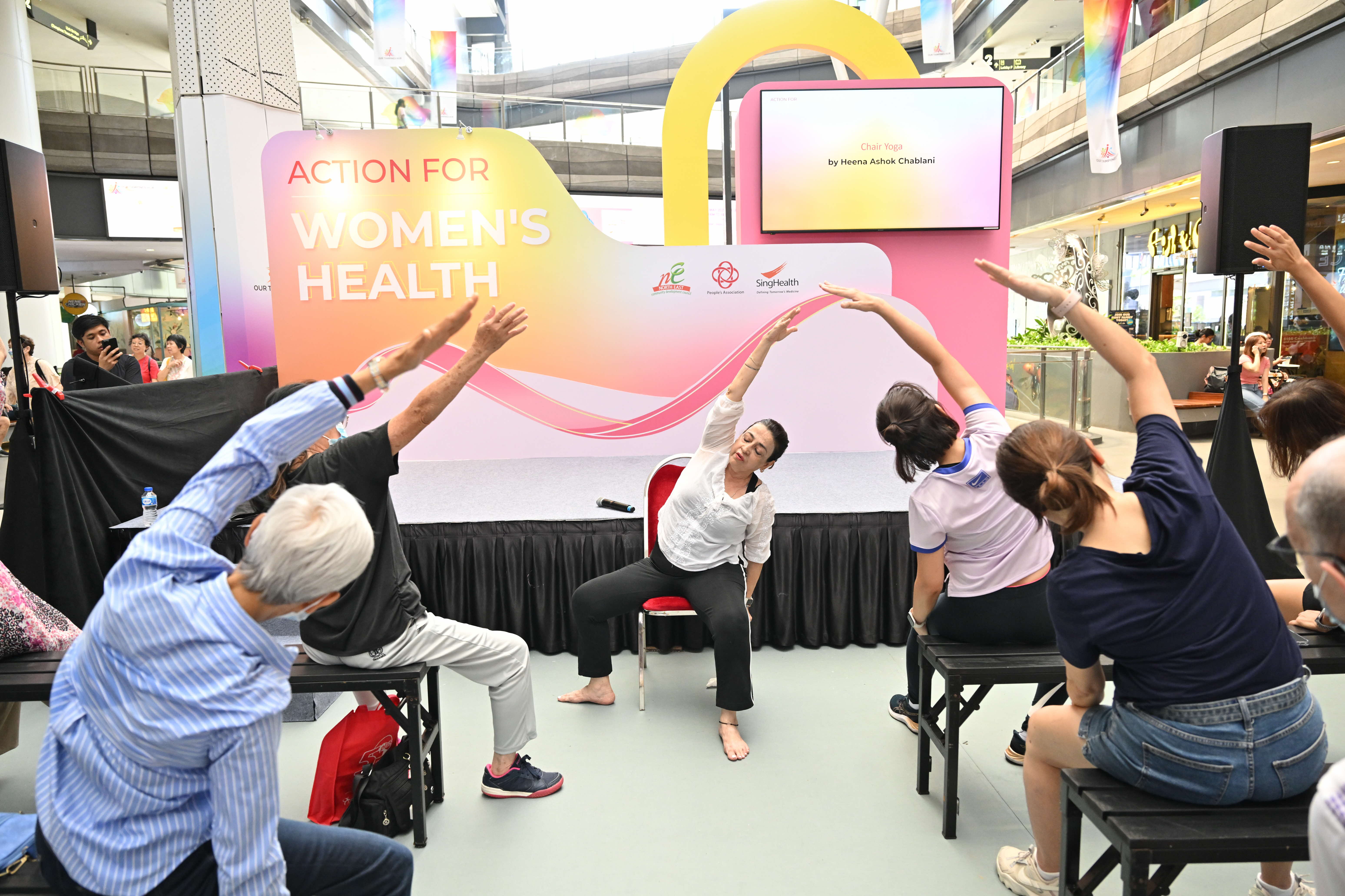 A group of people performing seated yoga stretches in front of a stage reading "Action for Women's Health."