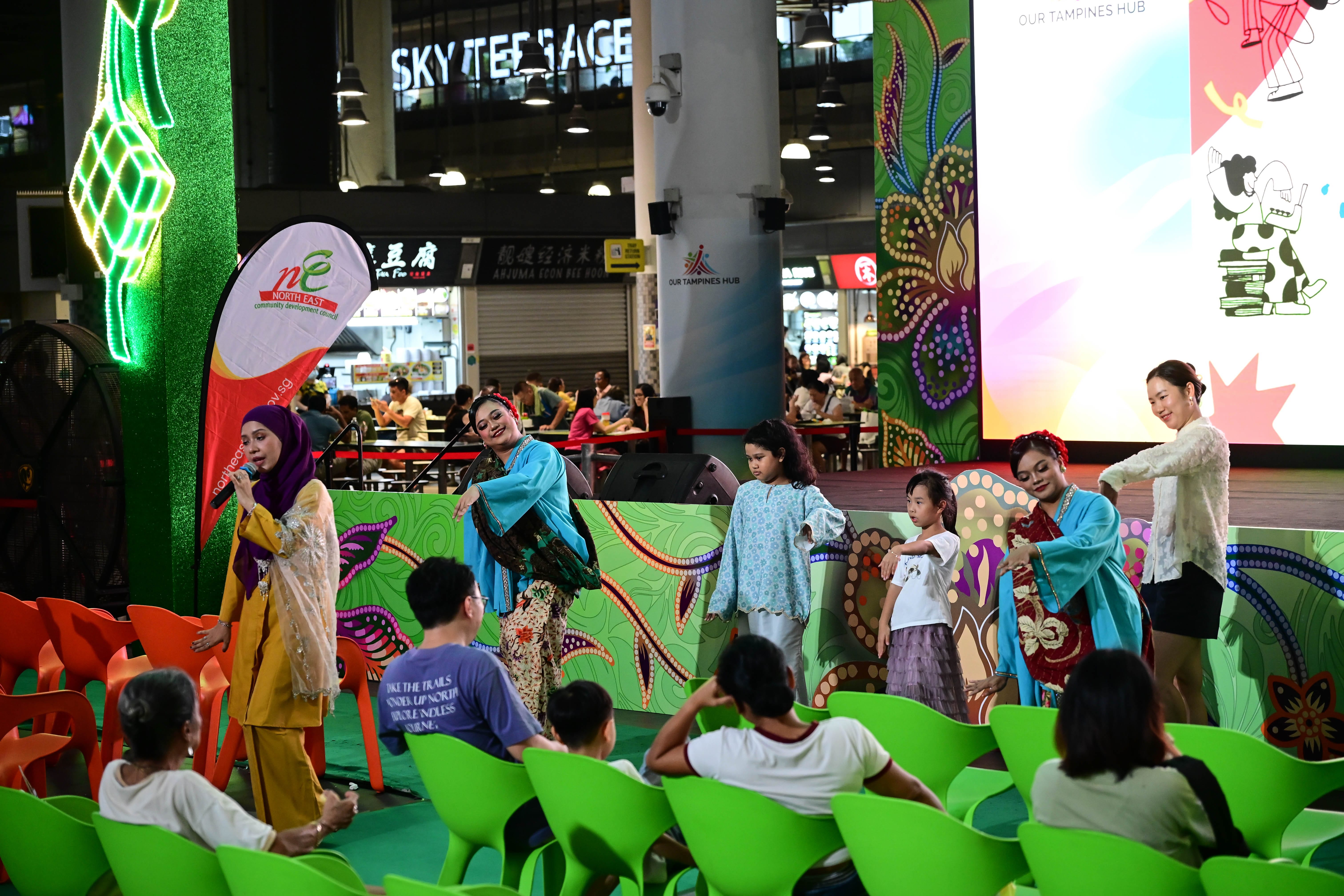 People wearing colourful clothes perform in front of a stage while an audience sits in green chairs watching them.