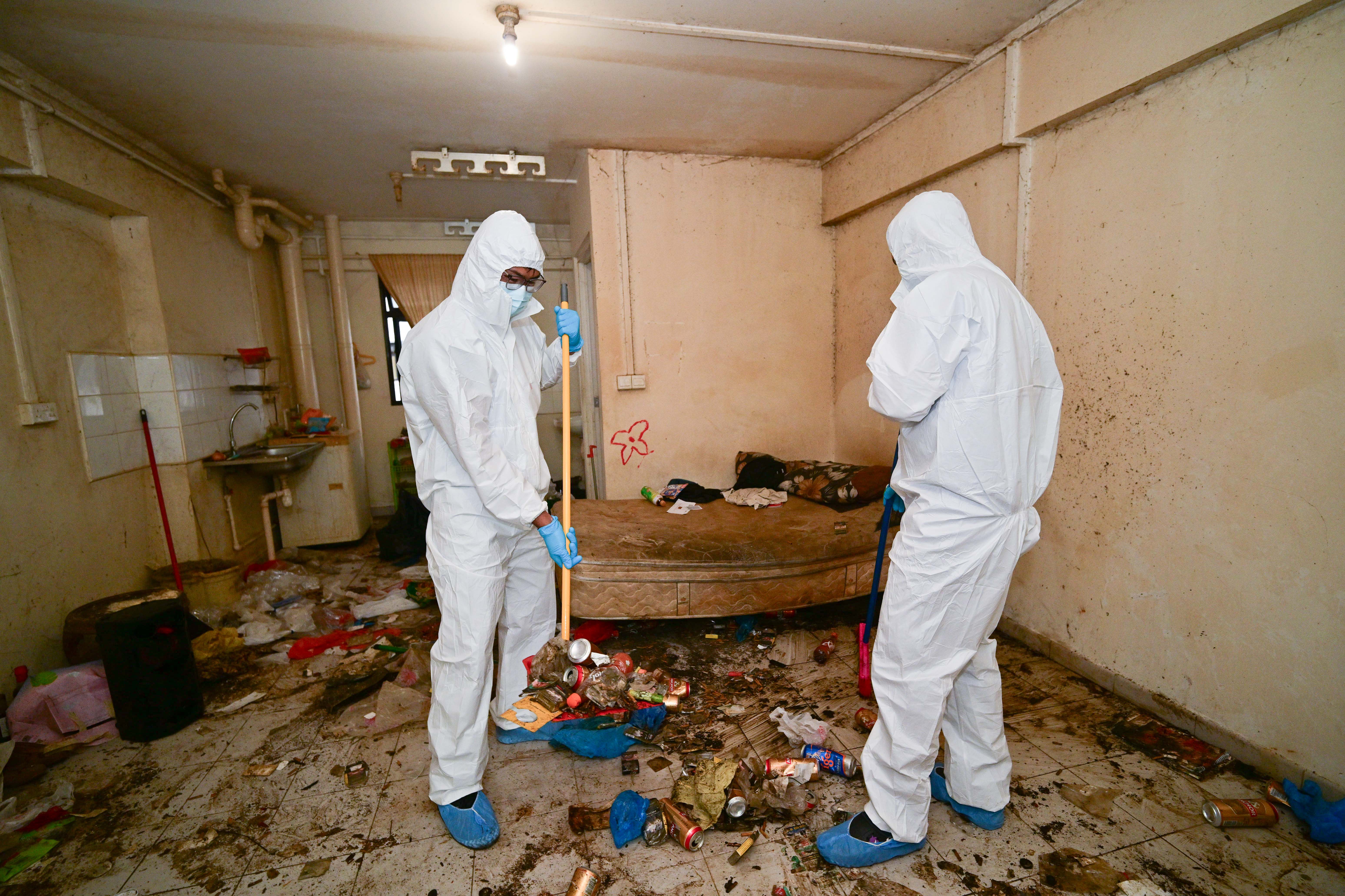 Two people in protective suits clean a dirty, messy room filled with trash and debris.