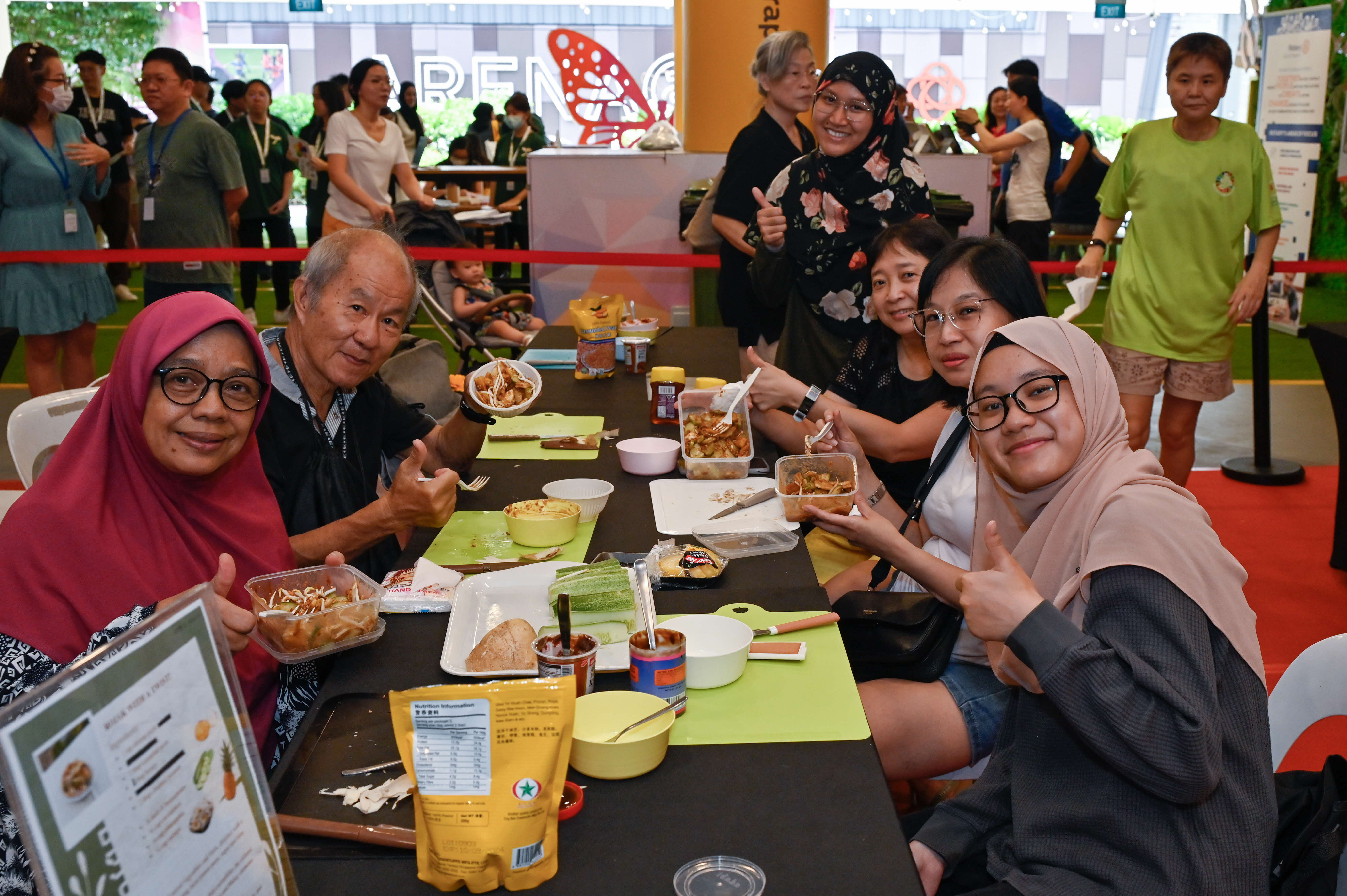 People seated at a table enjoying food, with more people standing and walking in the background under bright lighting.