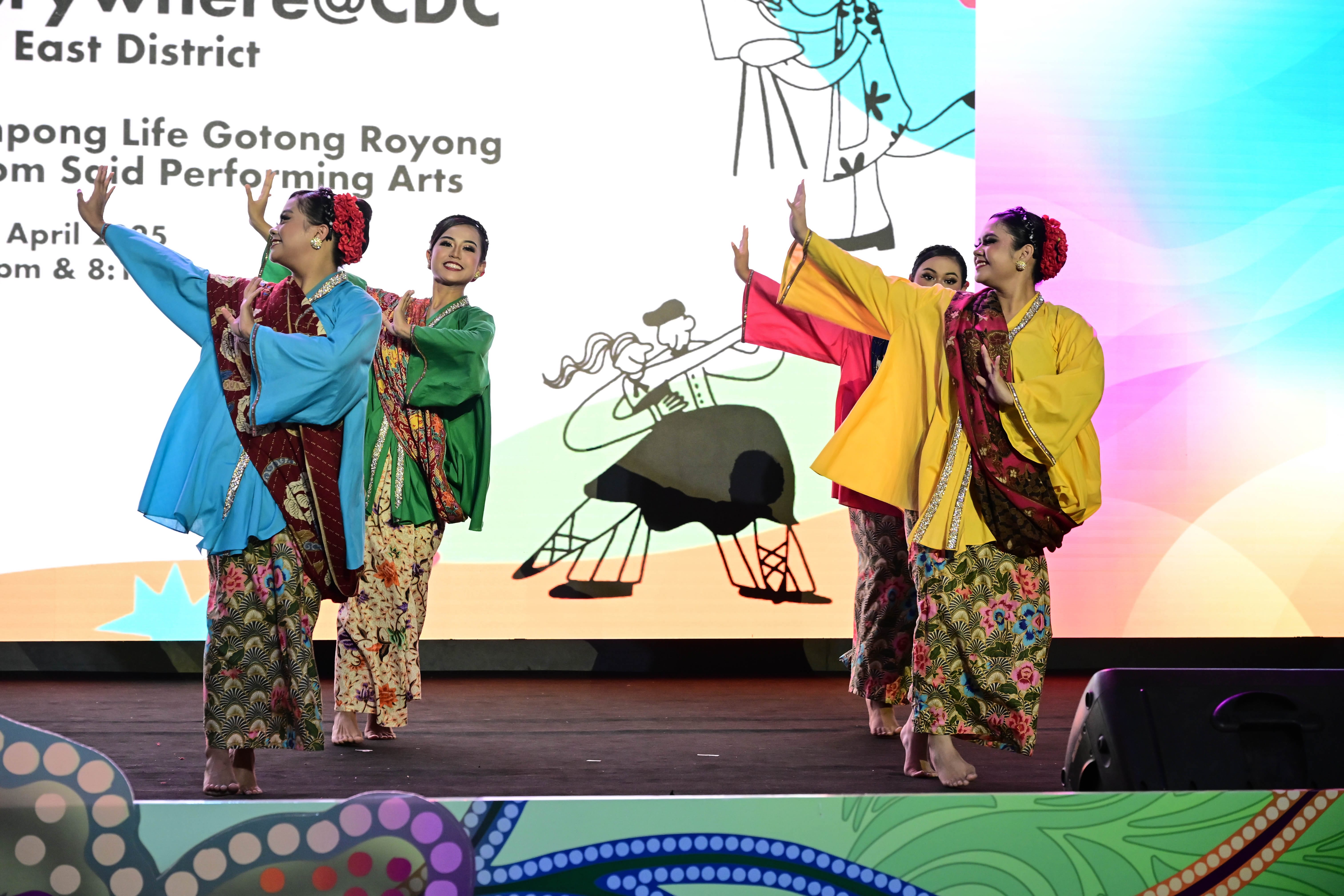 4 women perform a traditional dance in colourful costumes on stage with a graphic backdrop and text about an event.