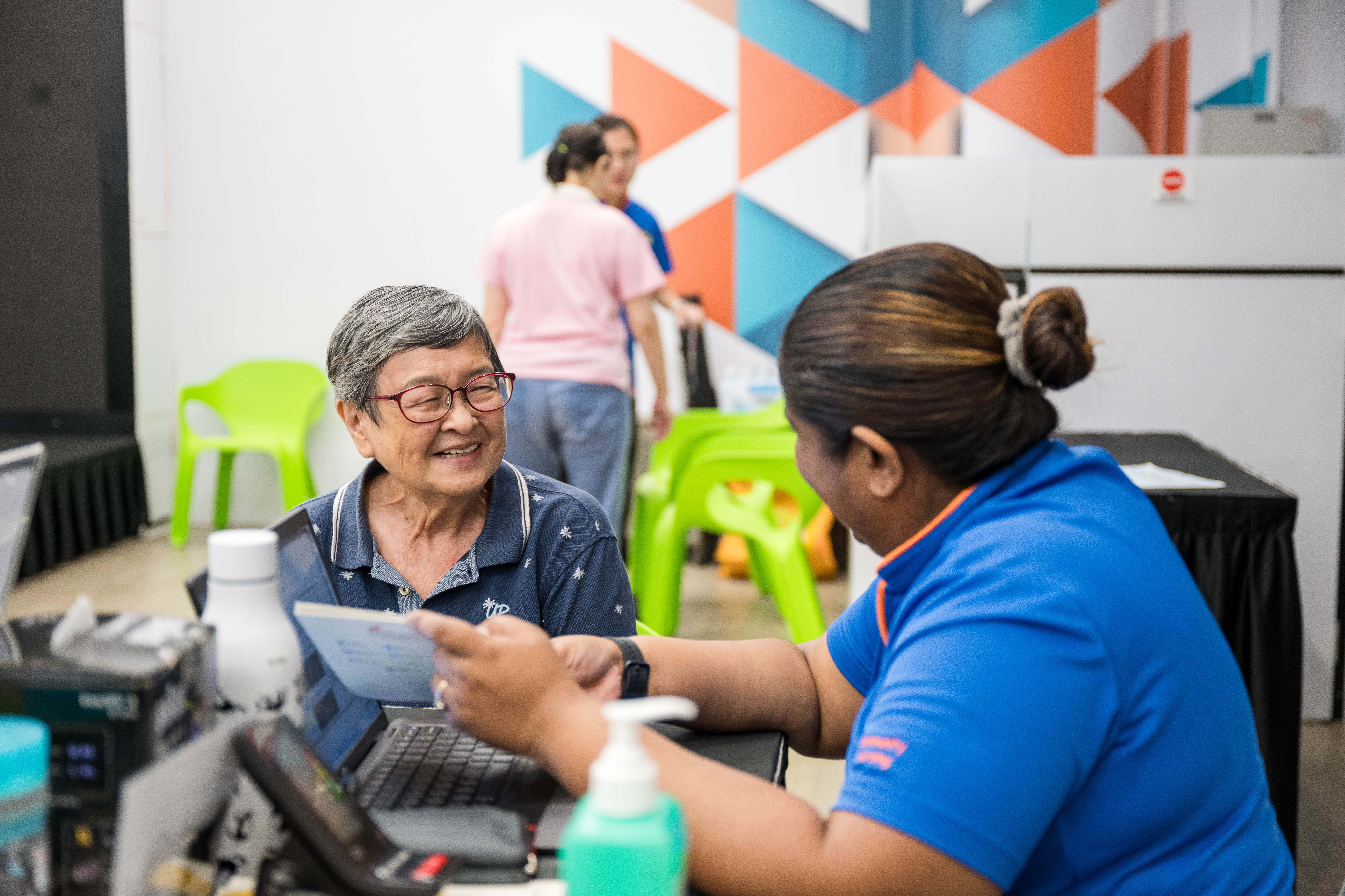 Two people are sitting at a table with a laptop and several documents in an indoor setting with colorful wall designs and green chairs.