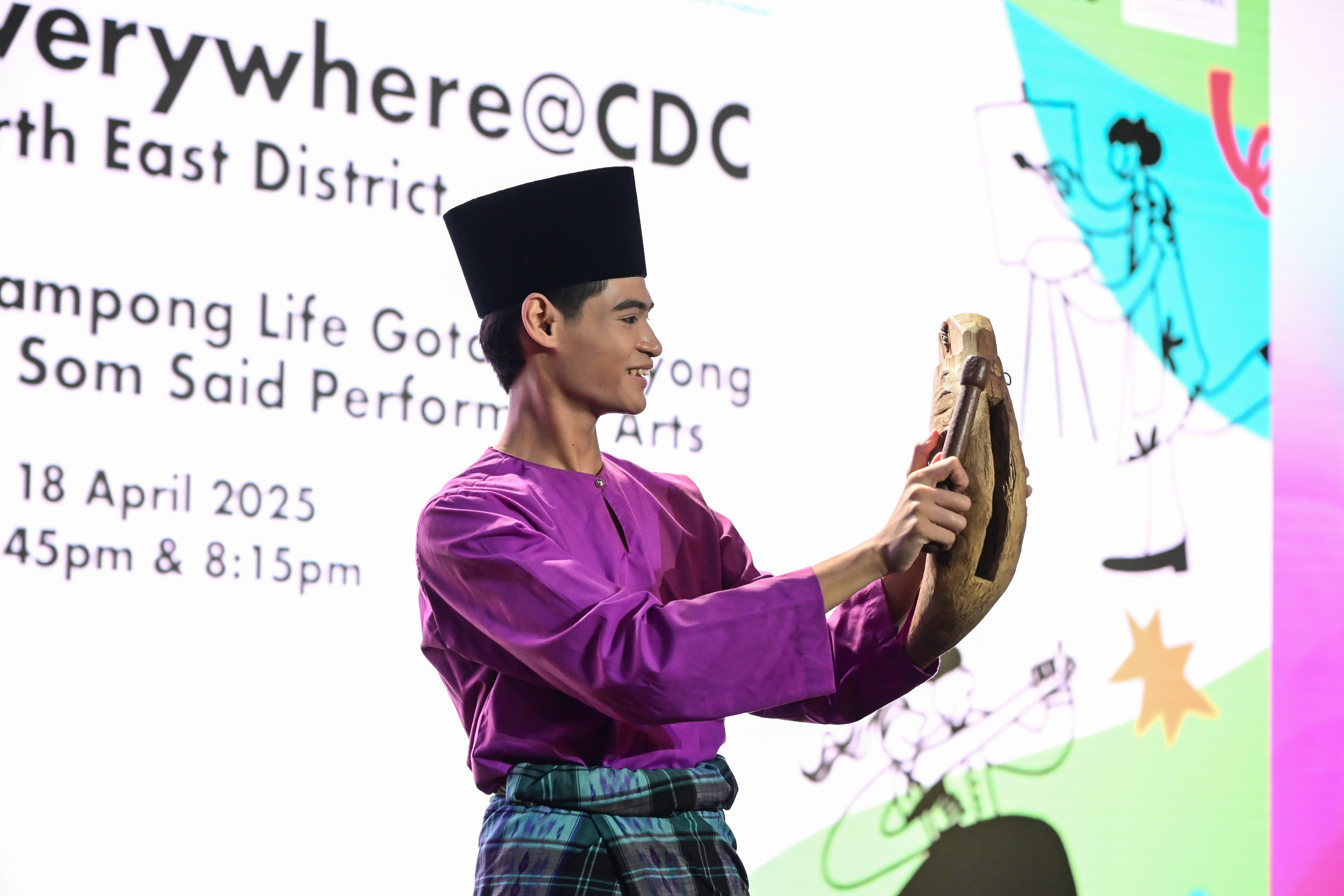 A person wearing traditional attire performs on stage while holding a wooden object; the backdrop displays event details.