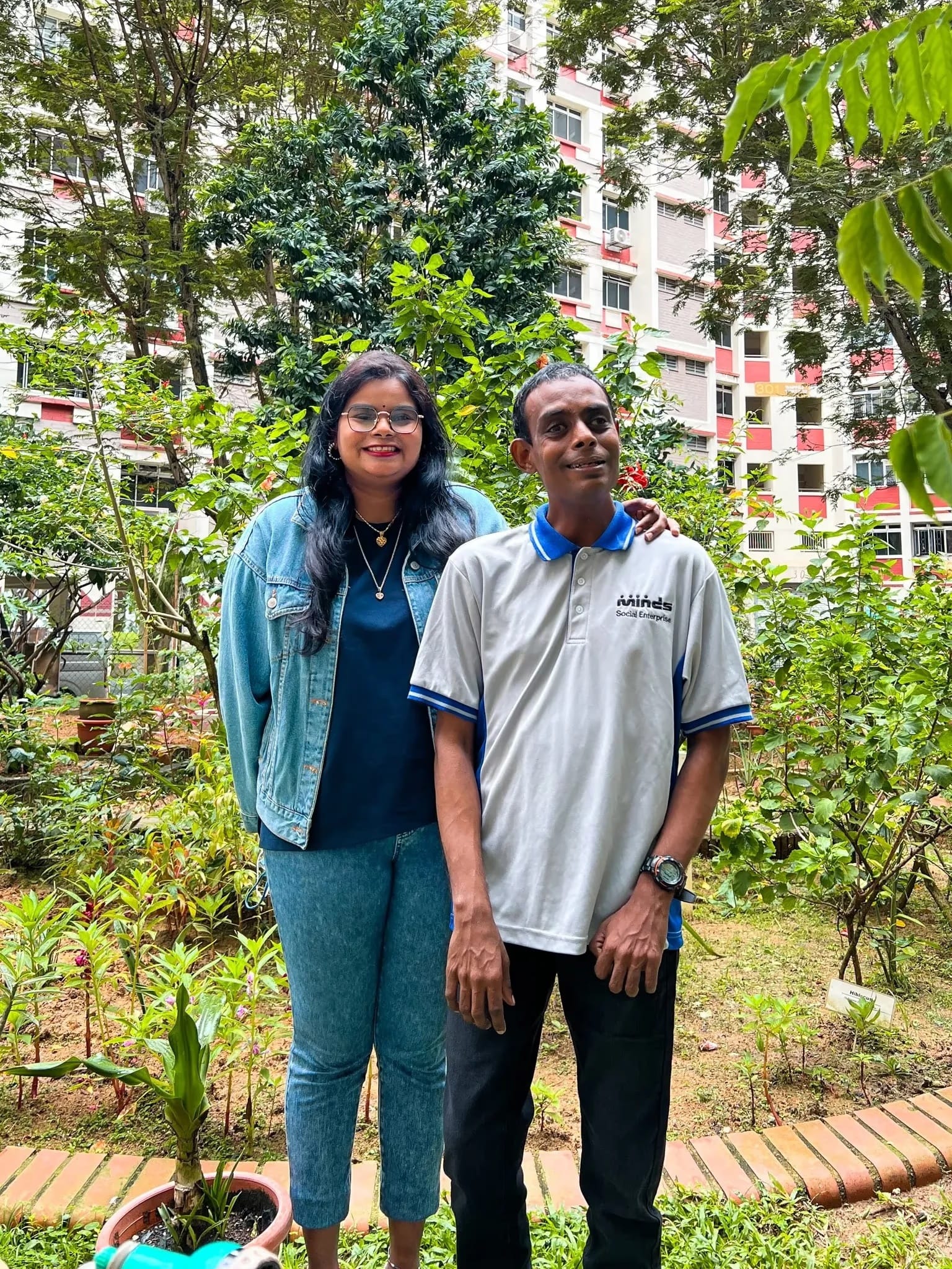 A man and a women stand outdoors beside each other among lush greenery and plants, with a residential building in the background.