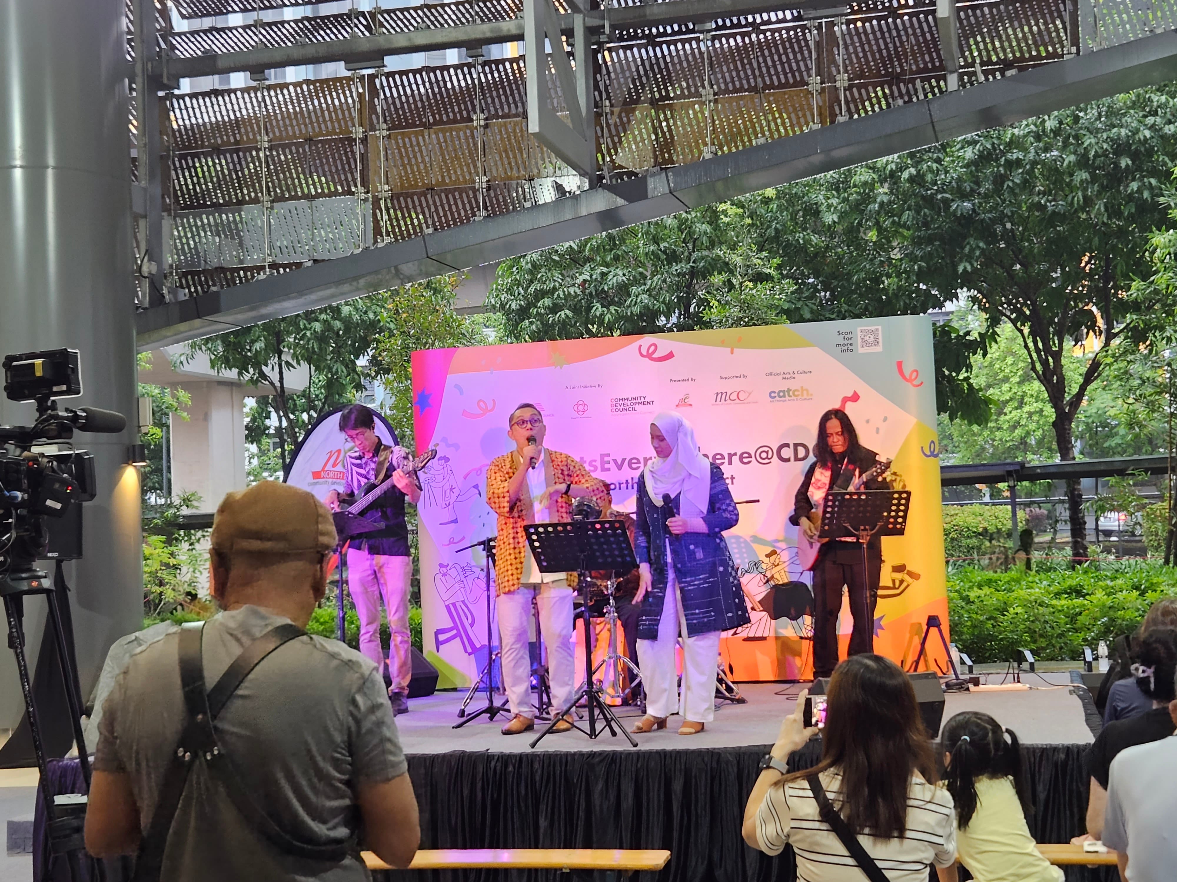 Live music performance at an outdoor community arts event, with a band and singers performing on stage in front of an audience.