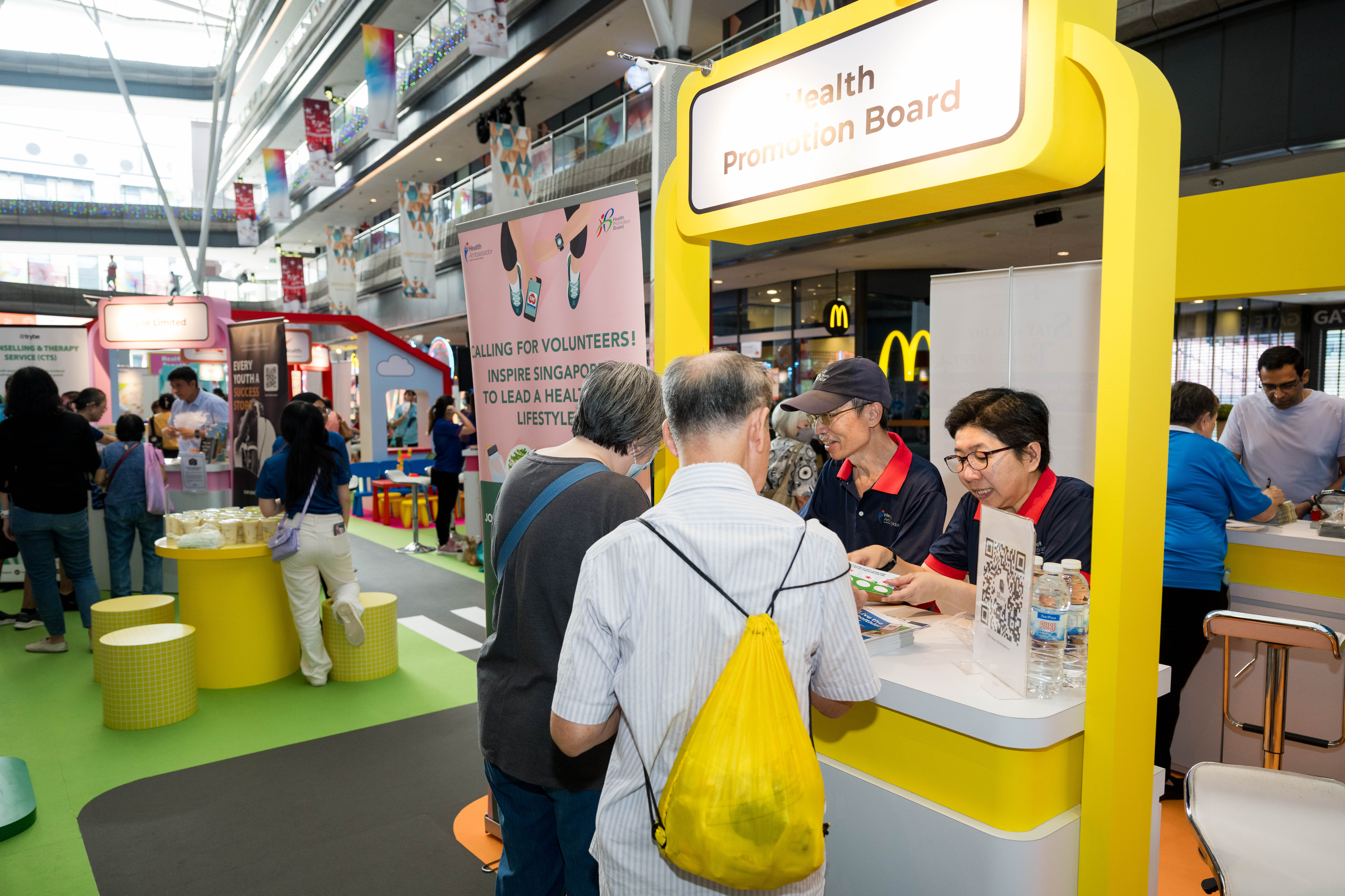 People interacting at the Health Promotion Board booth in a busy indoor public space.