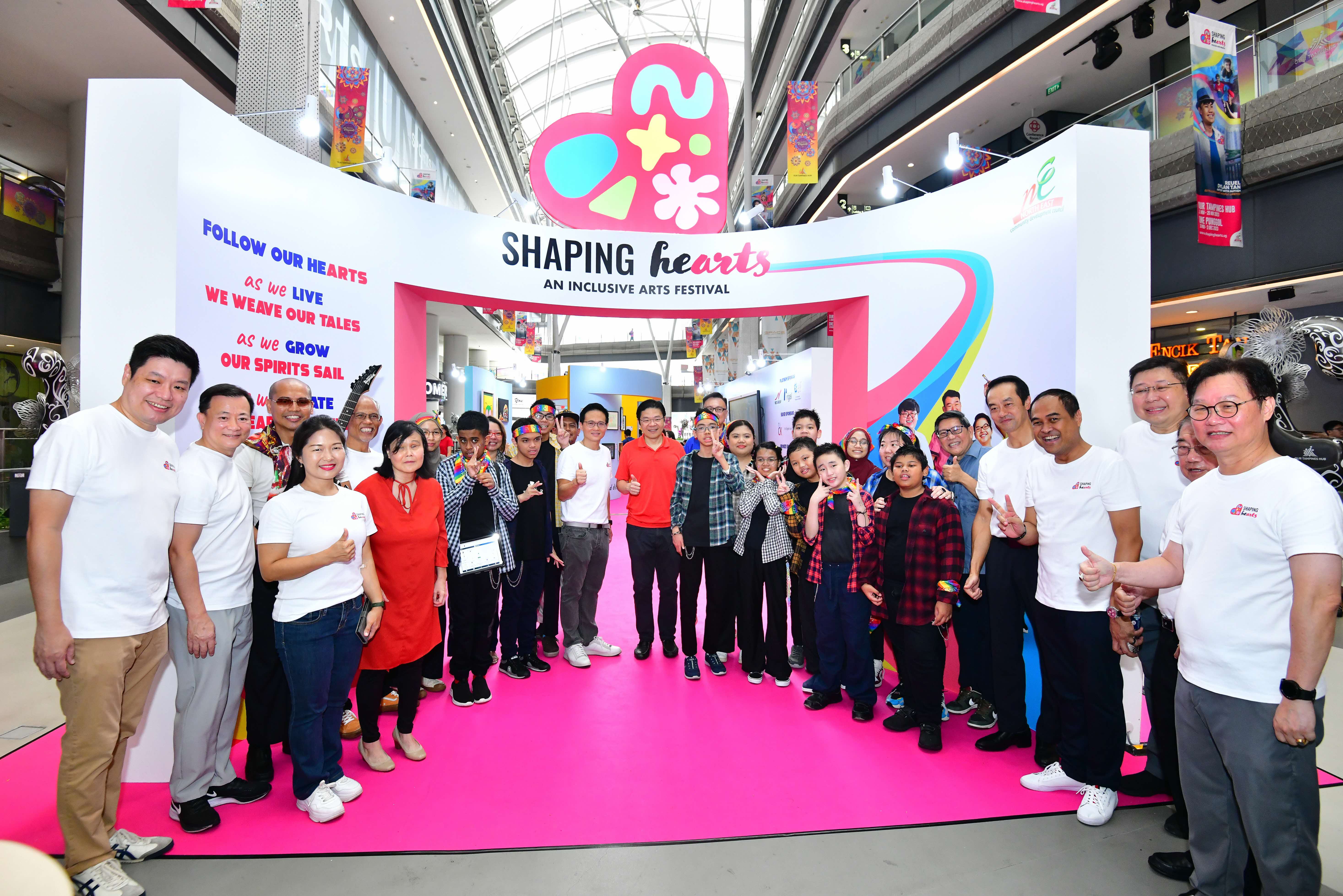 A group of people stand under a large, colorful sign for the Shaping Hearts Inclusive Arts Festival inside a bright, modern venue.