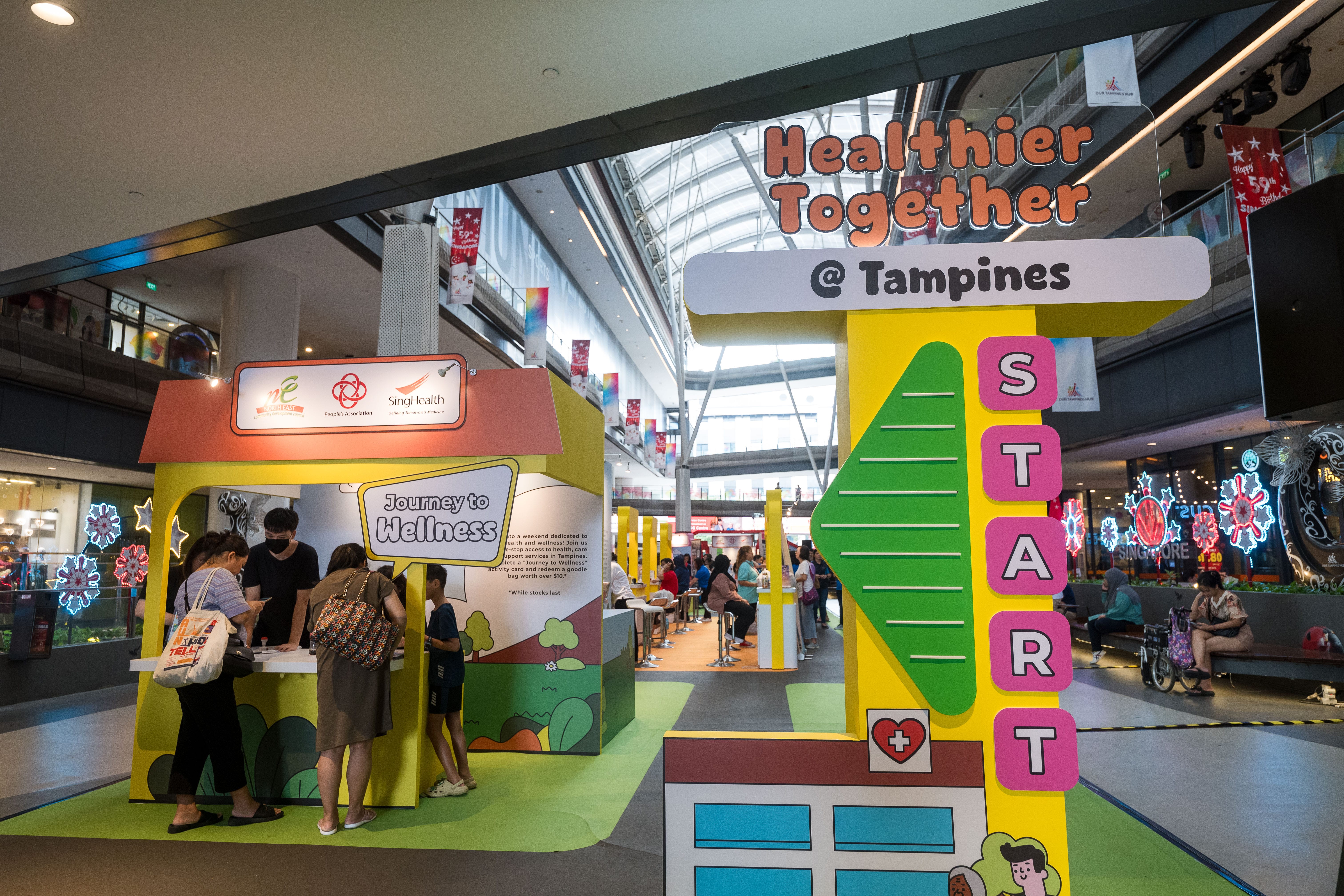 Indoor event with booths labeled "Journey to Wellness" and "Healthier Together @ Tampines" in a busy mall.