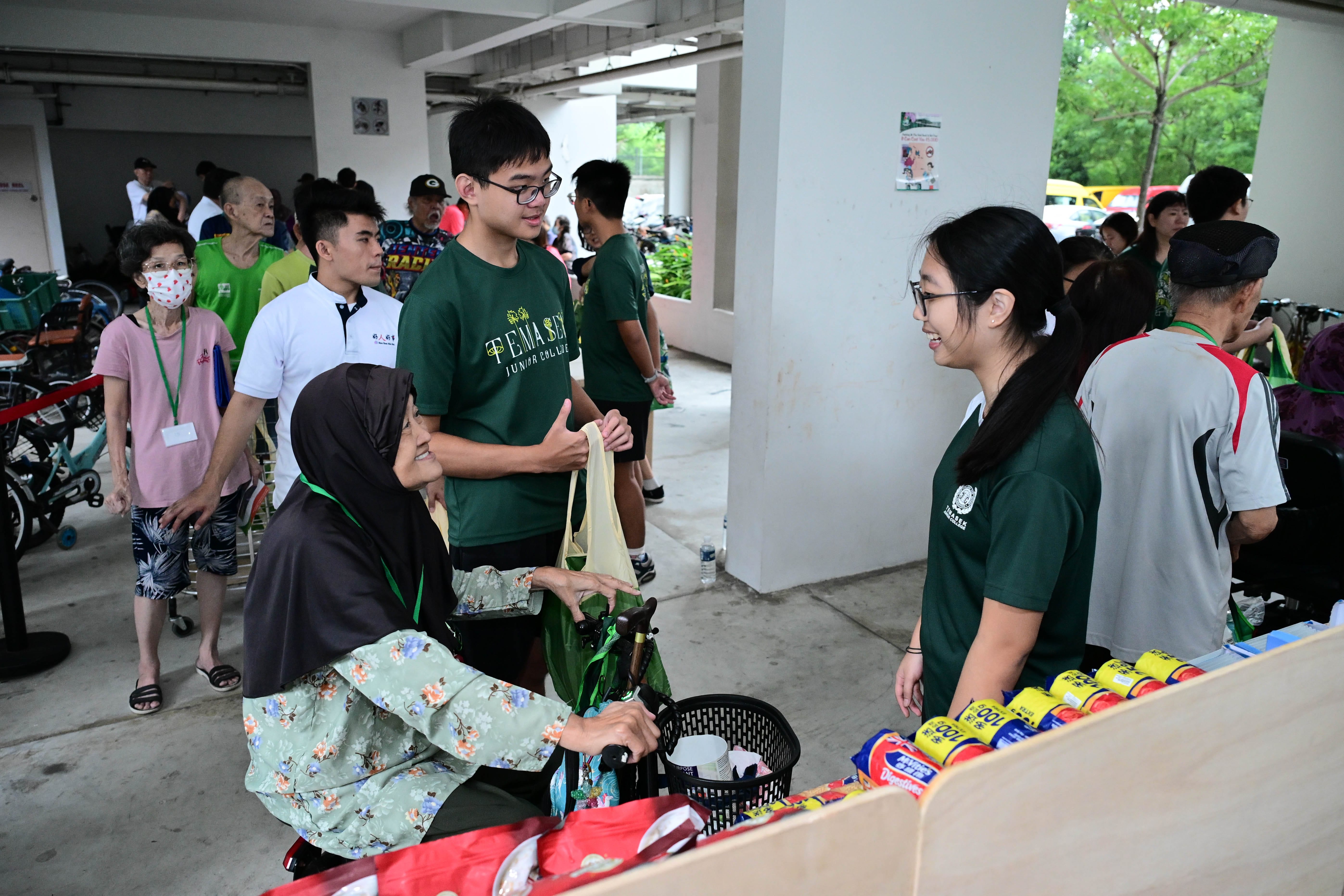 People are gathered at the void deck of a HDB, some in green shirts, with tables displaying various items in the foreground. An elderly lady in a PMD is talking to a lady in a green shirt