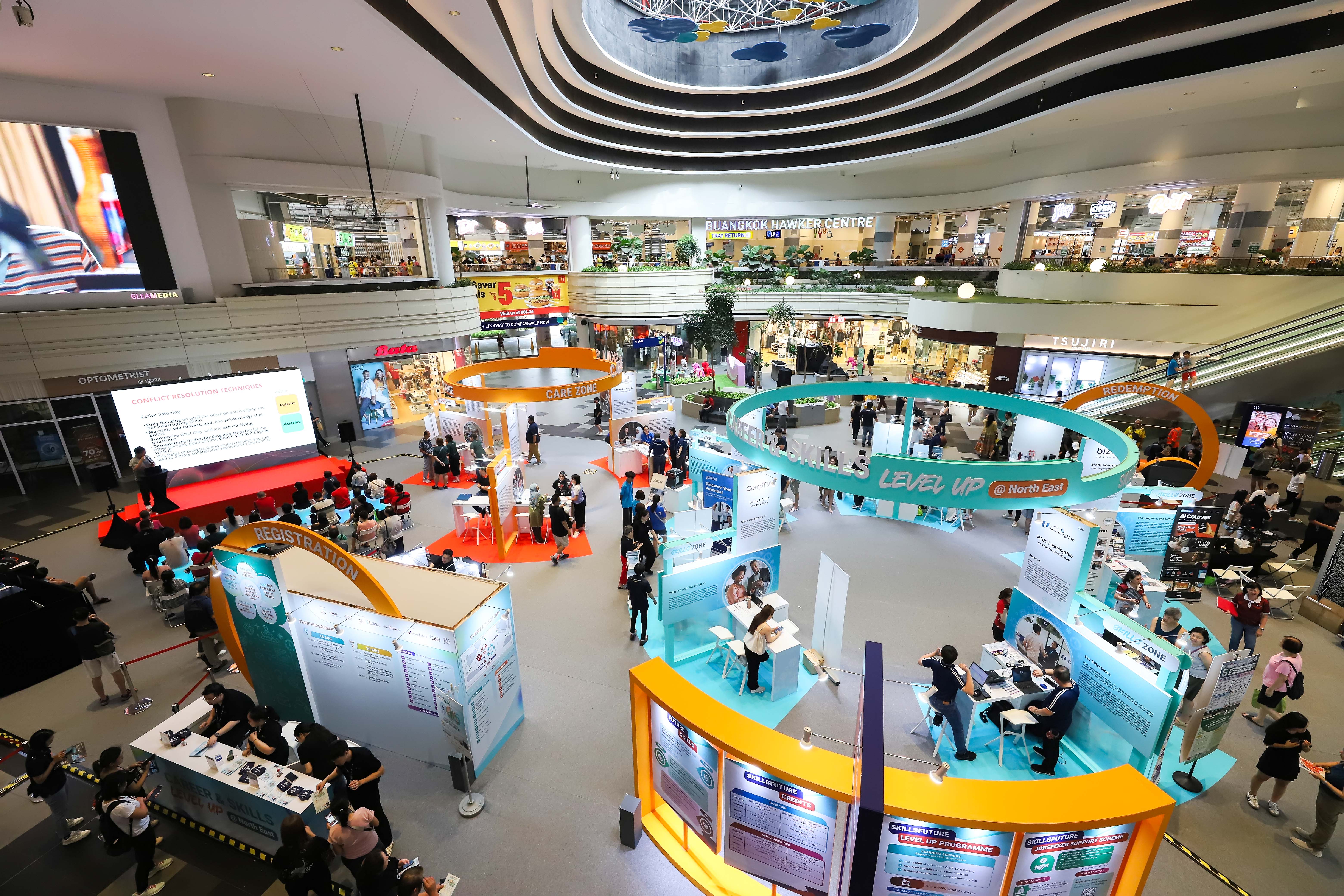 People exploring various educational booths at a career fair, with yellow, blue and orange decor and informational displays with a seating area and stage with a man giving a talk to an audience.