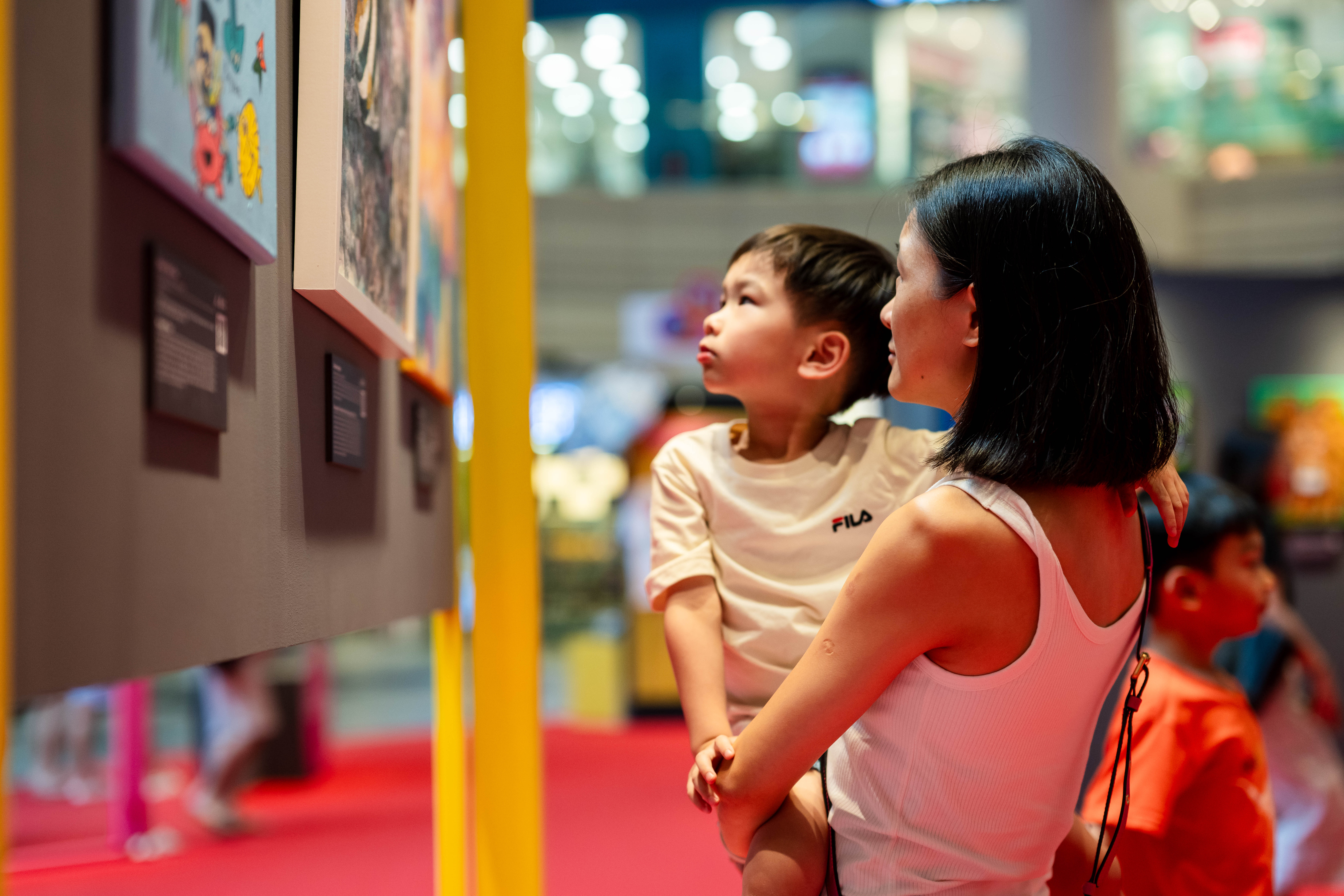 A woman holding a child is observing colorful artwork in a brightly lit gallery with red flooring.