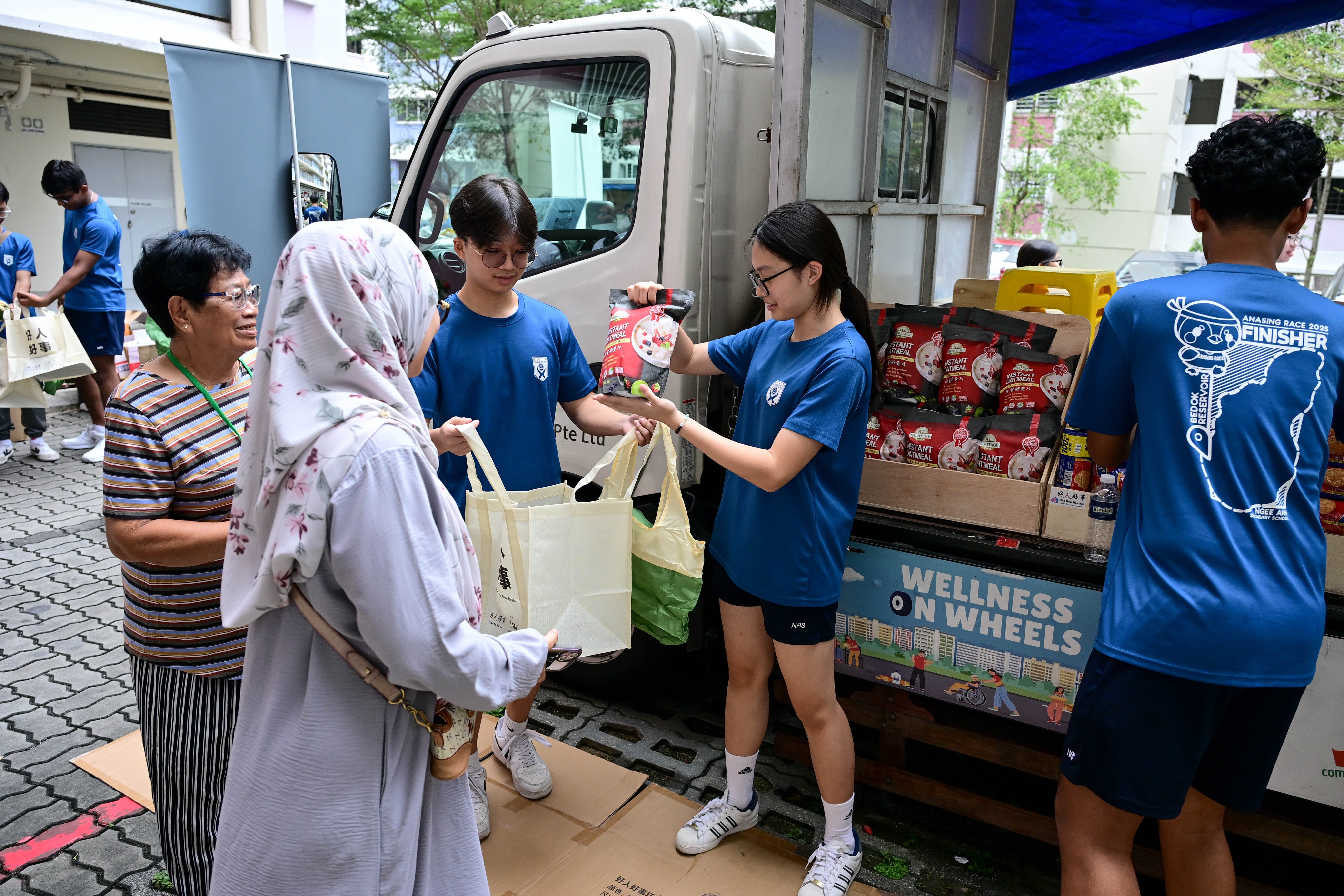 People in blue shirts handing out bags and food items from a truck with a "Wellness on Wheels" sign.