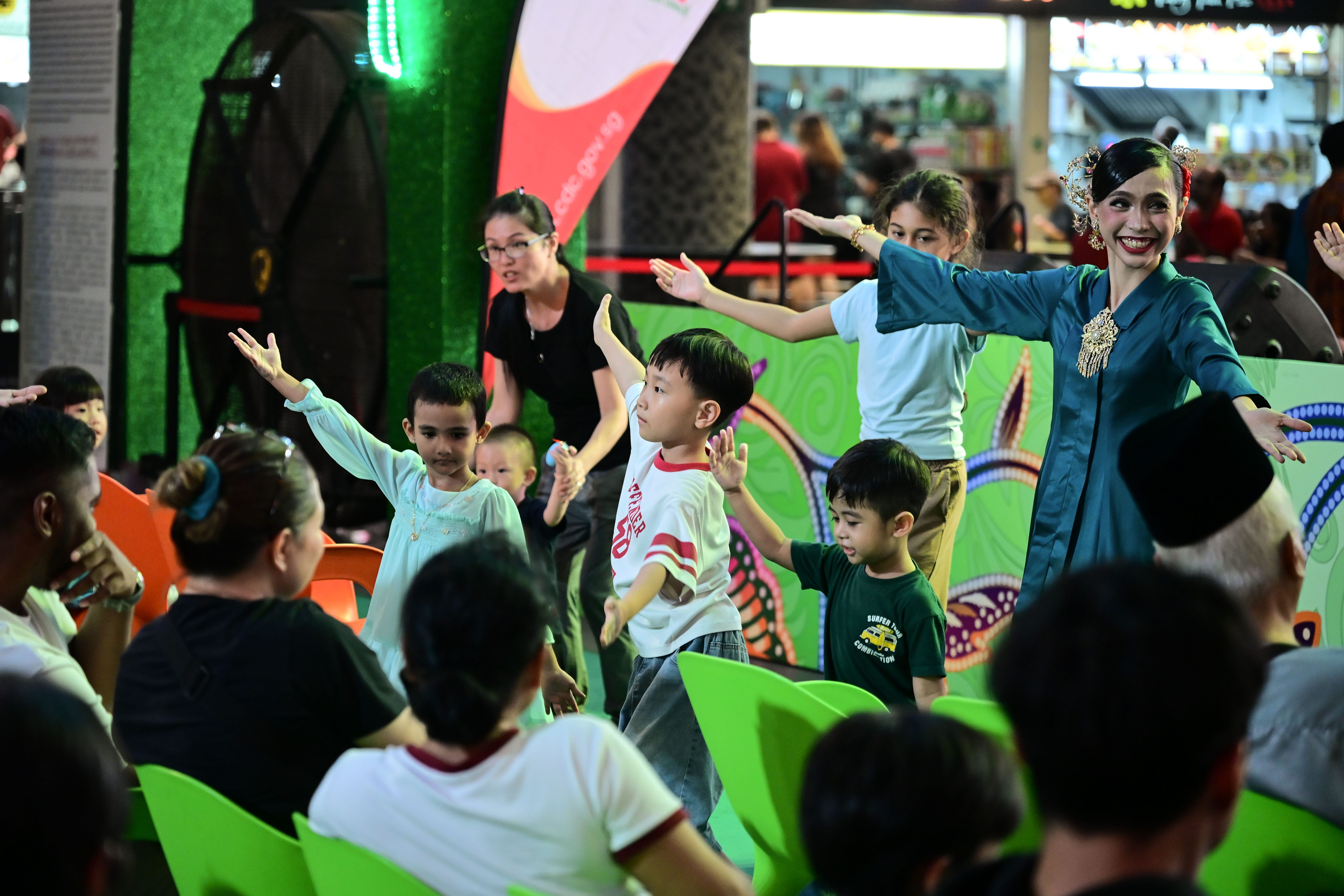 Children are standing and raising their arms, while adults sit and watch in a colorful, indoor setting.