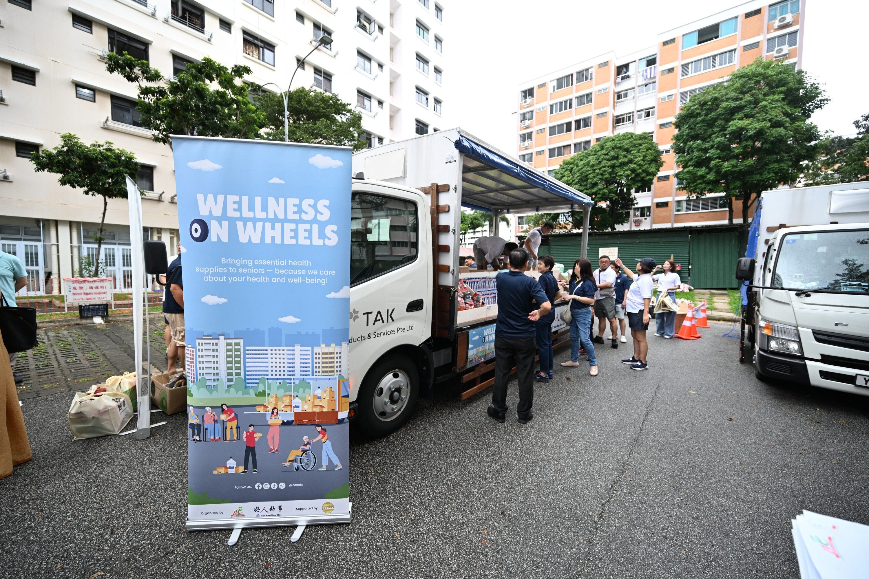 A banner reads "Wellness on Wheels" near a truck with people gathered around it in an outdoor urban area.