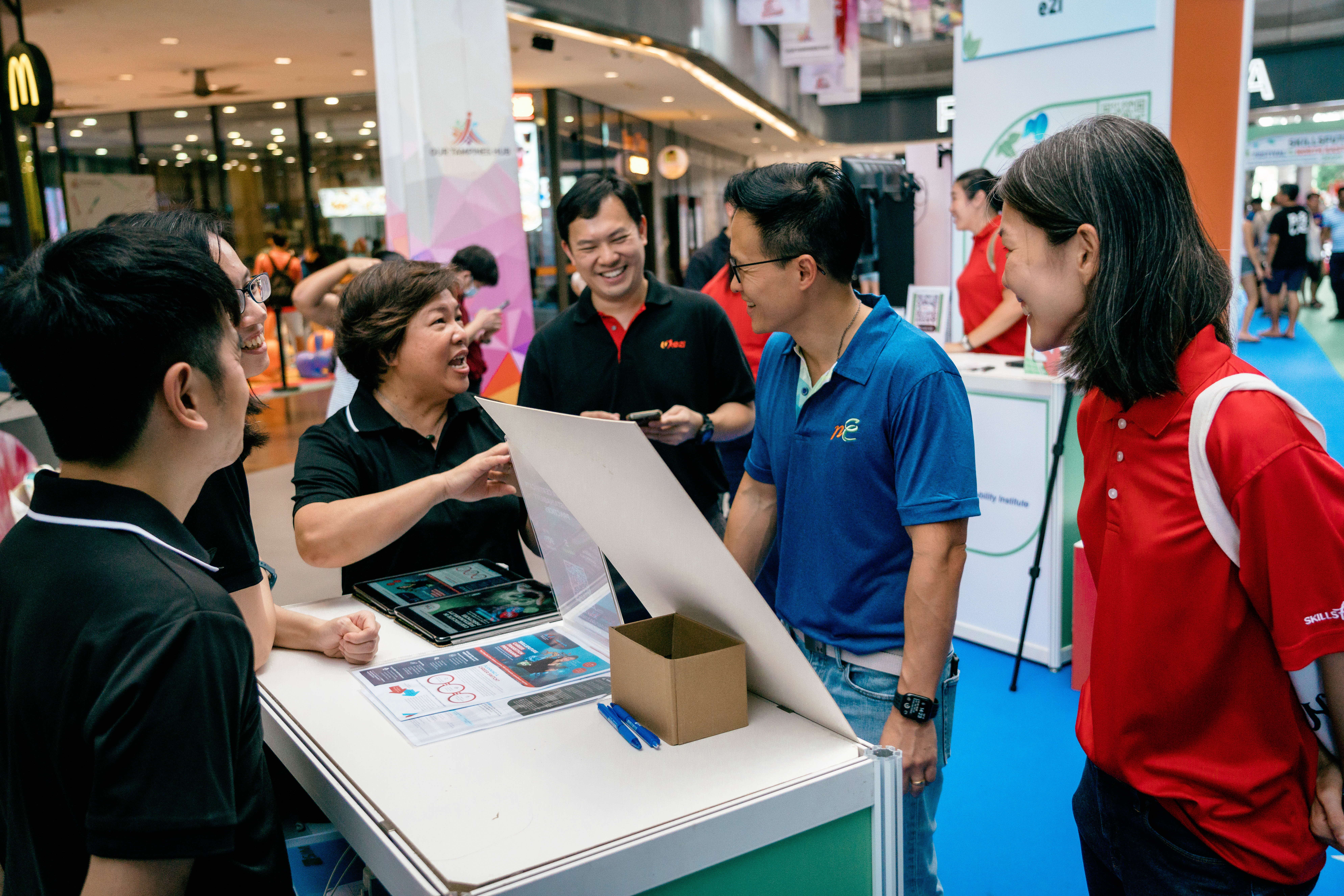 Several people are gathered around a booth in a busy indoor event space, talking and looking at materials on a table.