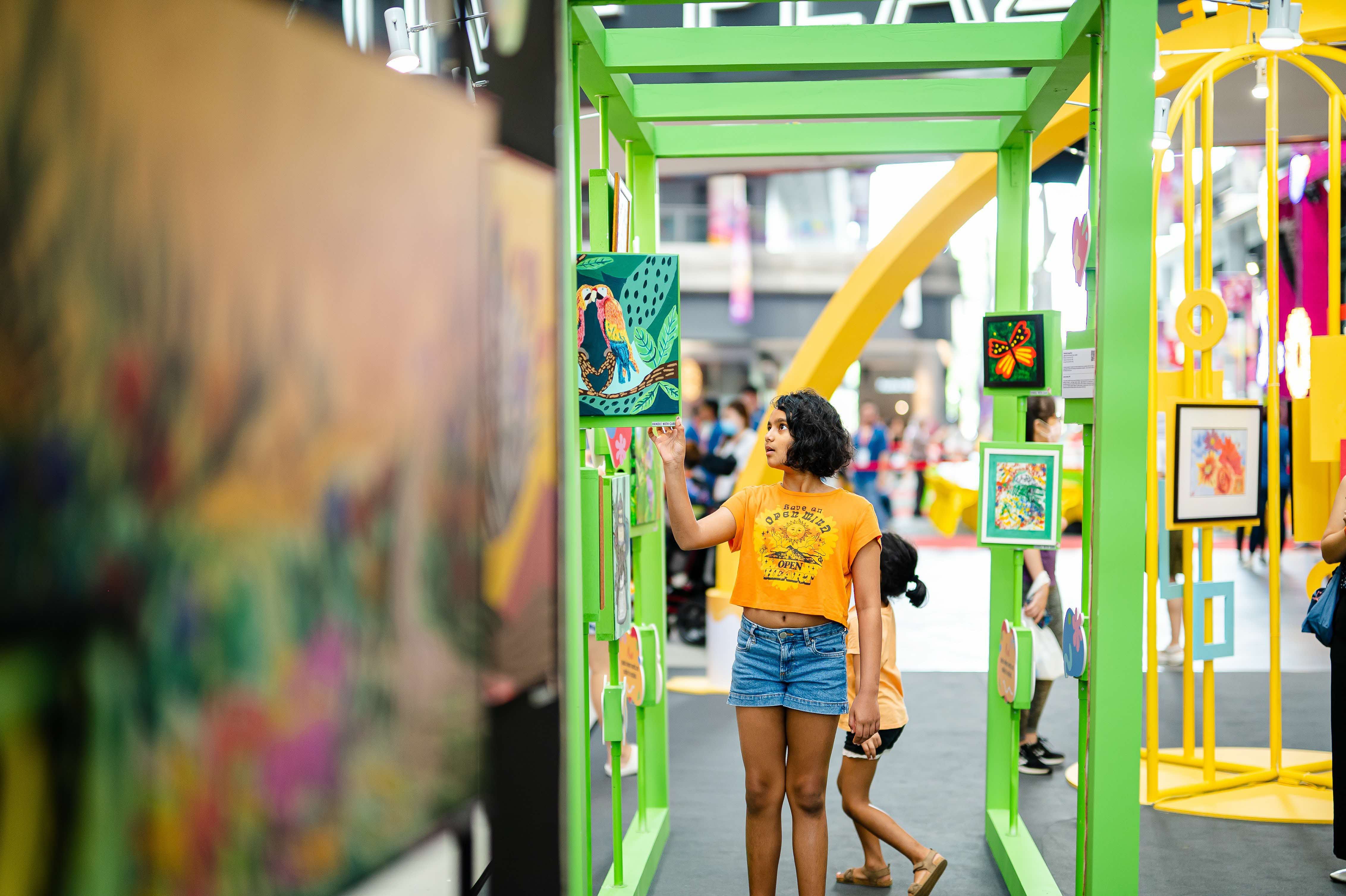People interacting with colourful artwork displayed on green frames at an indoor art exhibit.